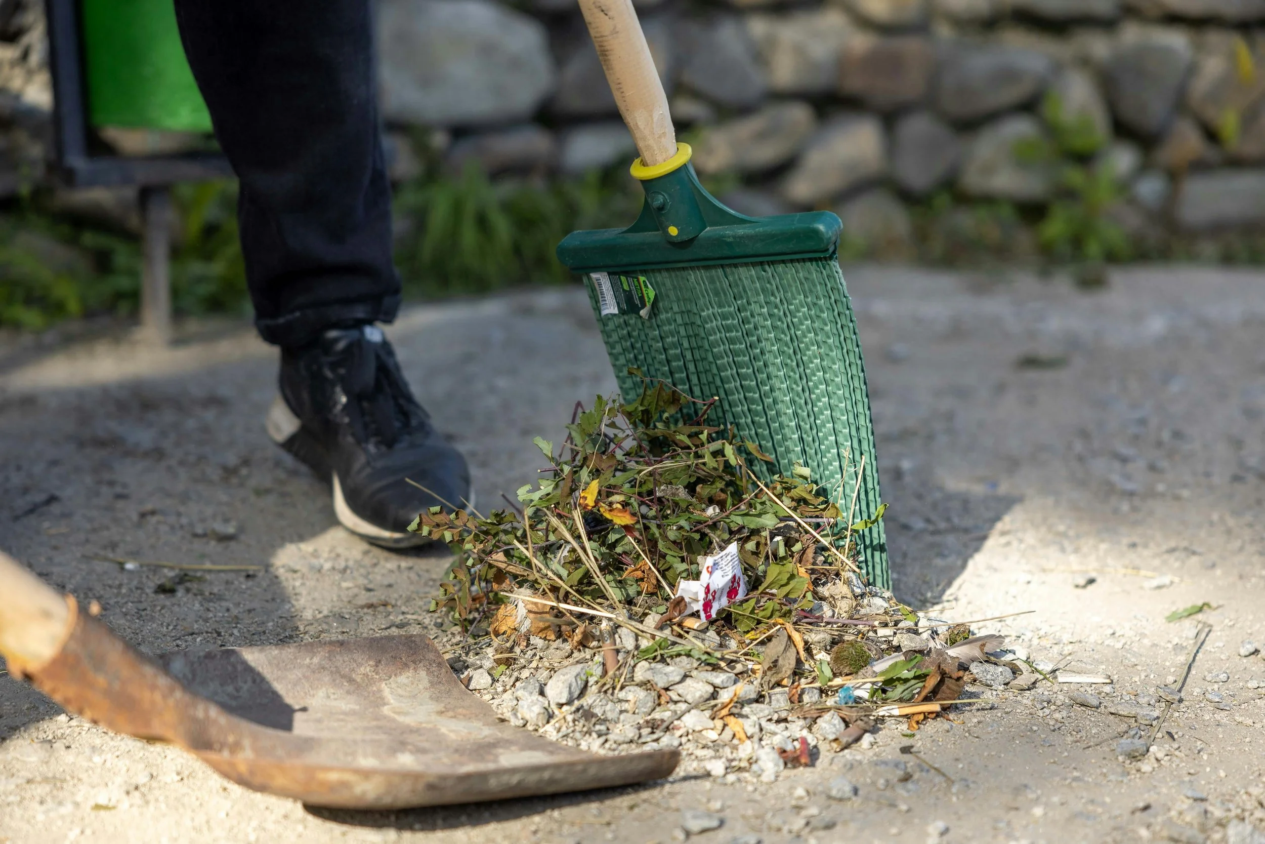 Person shoveling trash and leaves from the ground using a green dustpan and a hoe, with a stone wall in the background.