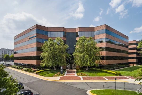 Modern multi-story office building with brick and reflective glass facade, landscaped lawn, trees, and paved entrance under a partly cloudy sky