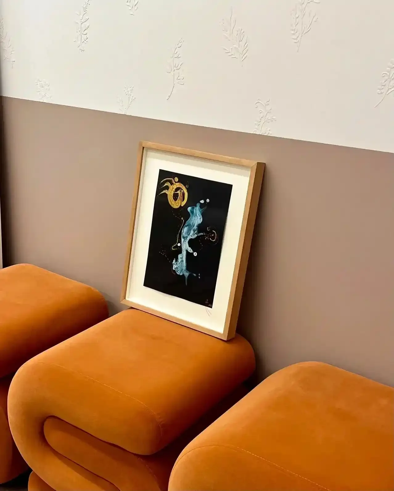 An indoor waiting area with orange cushioned chairs, a framed abstract artwork leaning against the wall, and patterned wallpaper with leaf designs.
