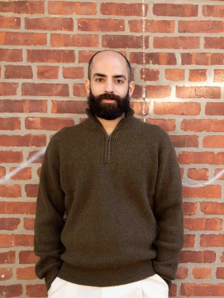Man with a beard and bald head standing in front of a red brick wall, wearing a brown sweater and white pants.