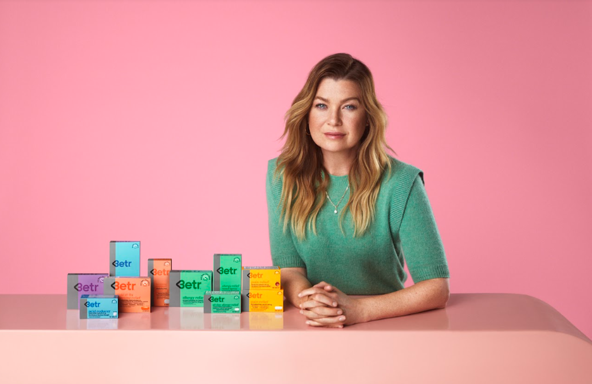 Actor, activist and BETR remedies co-founder, Ellen Pompeo, wearing a green sweater, stands behind a counter with various boxes of "BETR" branded medicine on a pink background.