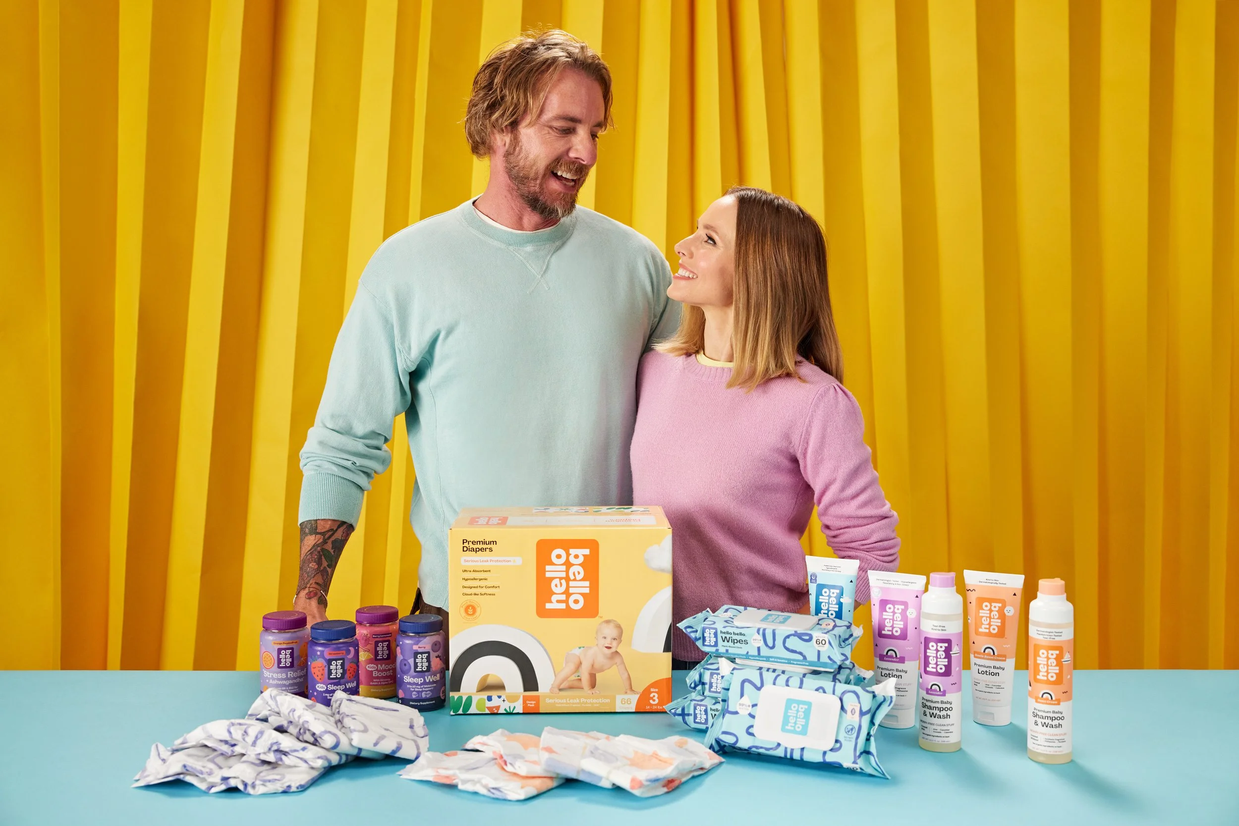 Two people standing behind a table with Hello Bello products, including diapers, wipes, lotions, and bottles, in front of a yellow background.