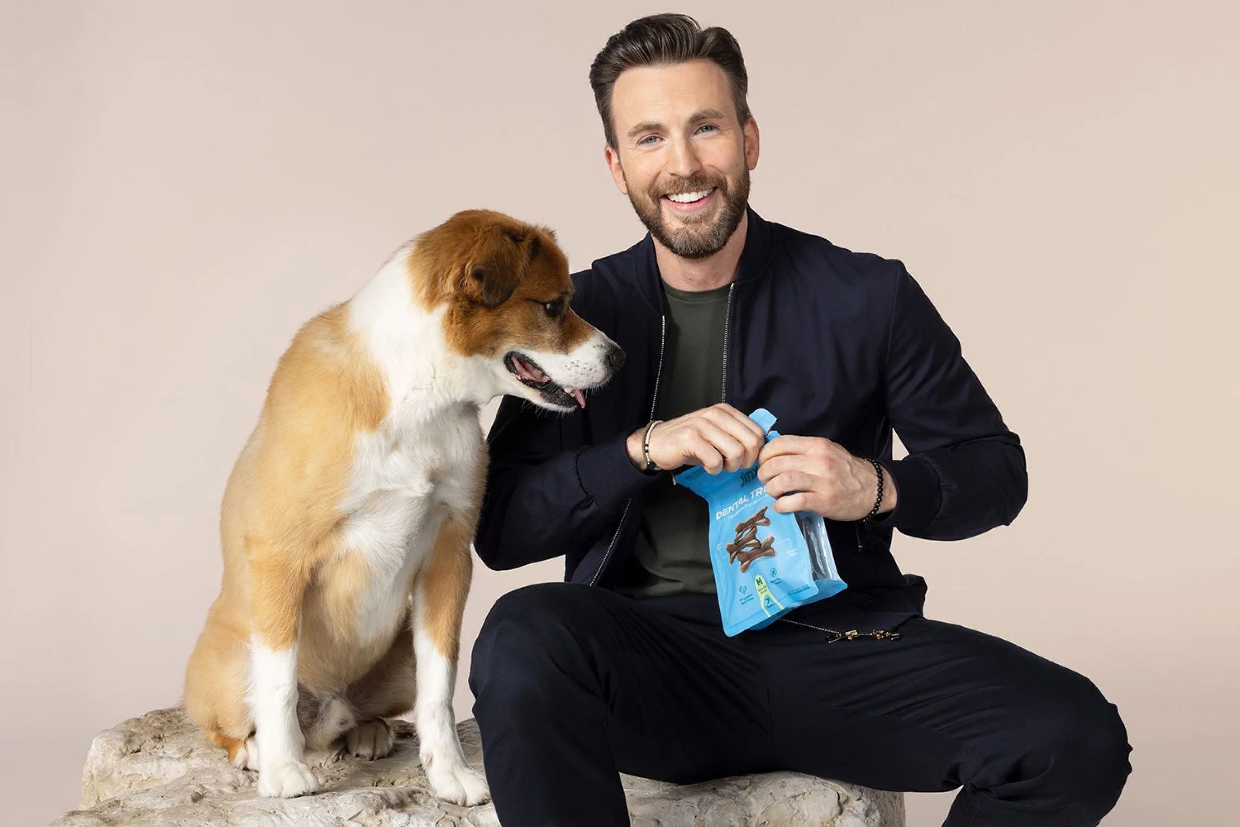 Actor, dog dad and Jinx co-founder, Chris Evans, holding dog dental treats with his dog sitting beside him on a stone surface against a neutral background.