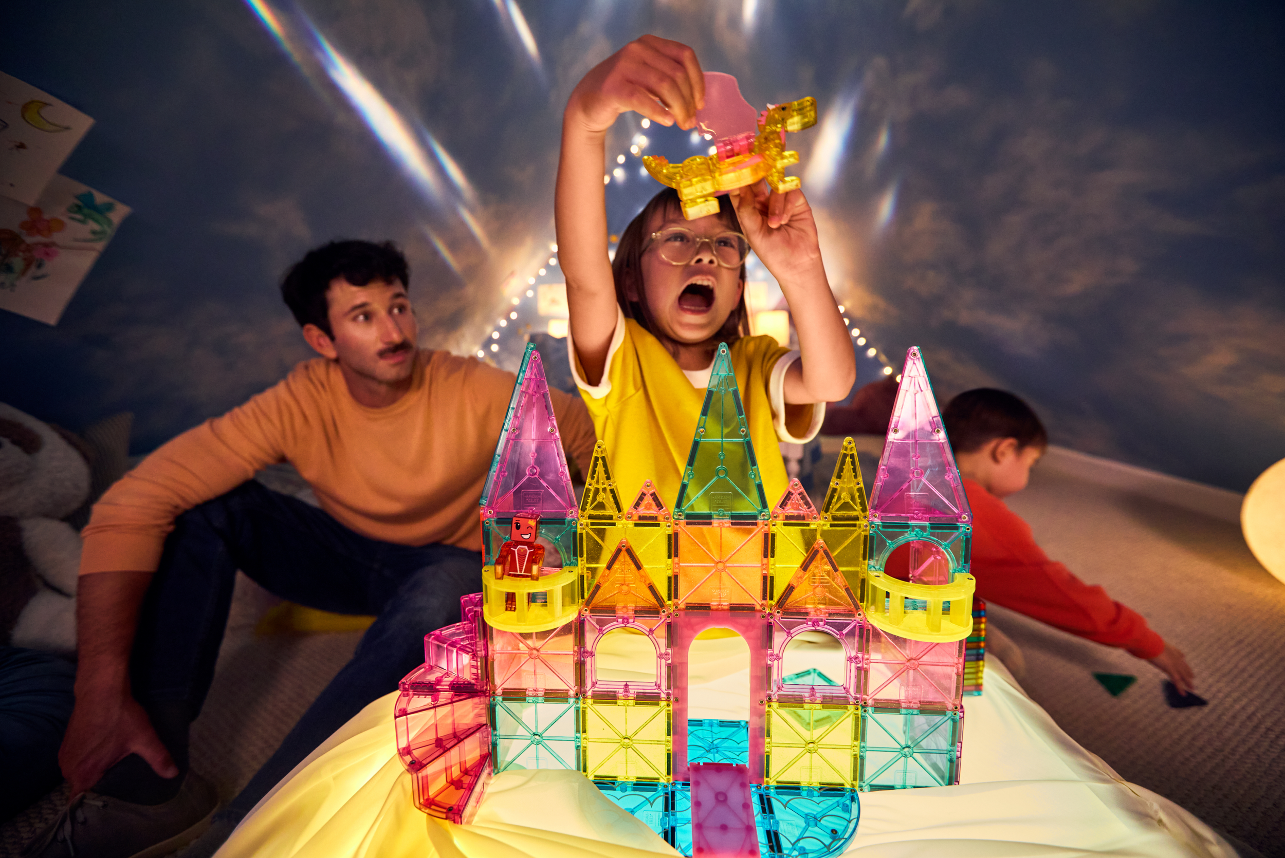 Child playing with colorful magnetic building tiles, assembling a castle structure, while an adult observes and another child plays in the background, in a cozy, decorated room.
