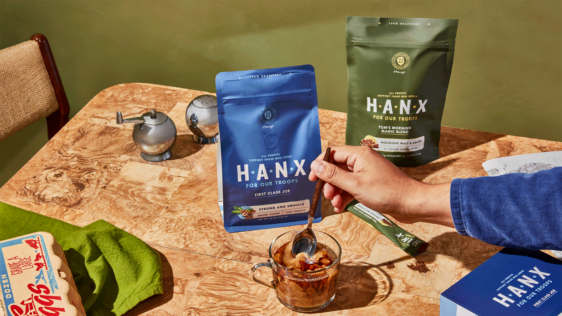 Person stirring coffee with HANX For Our Troops coffee packs on a table, alongside vintage salt and pepper shakers, an egg carton, and a green napkin.
