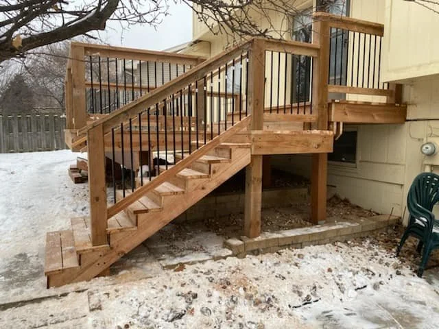 Wooden stairs and deck extending from a house, with a black metal railing, in a snowy backyard.
