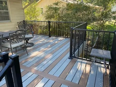 Outdoor deck with metal railing, outdoor table with chairs, and a outdoor chair on the right side, surrounded by trees and greenery.