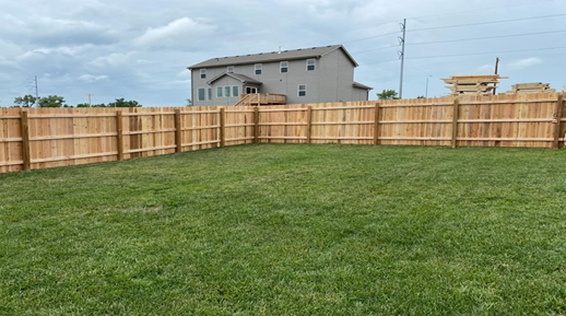 A backyard with a green lawn, a wooden privacy fence, and a house in the background under a cloudy sky.