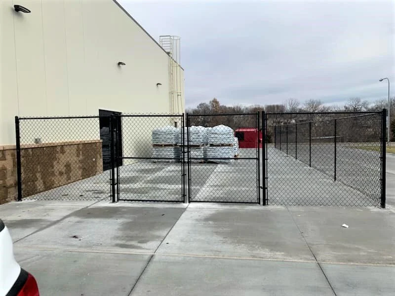 A fenced area with three large white wrapped pallets behind a black chain-link gate, adjacent to a beige building, with a red mailbox and a parking lot.