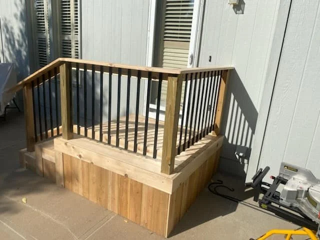 Wooden deck with railing installed on a concrete patio beside a house with gray siding.
