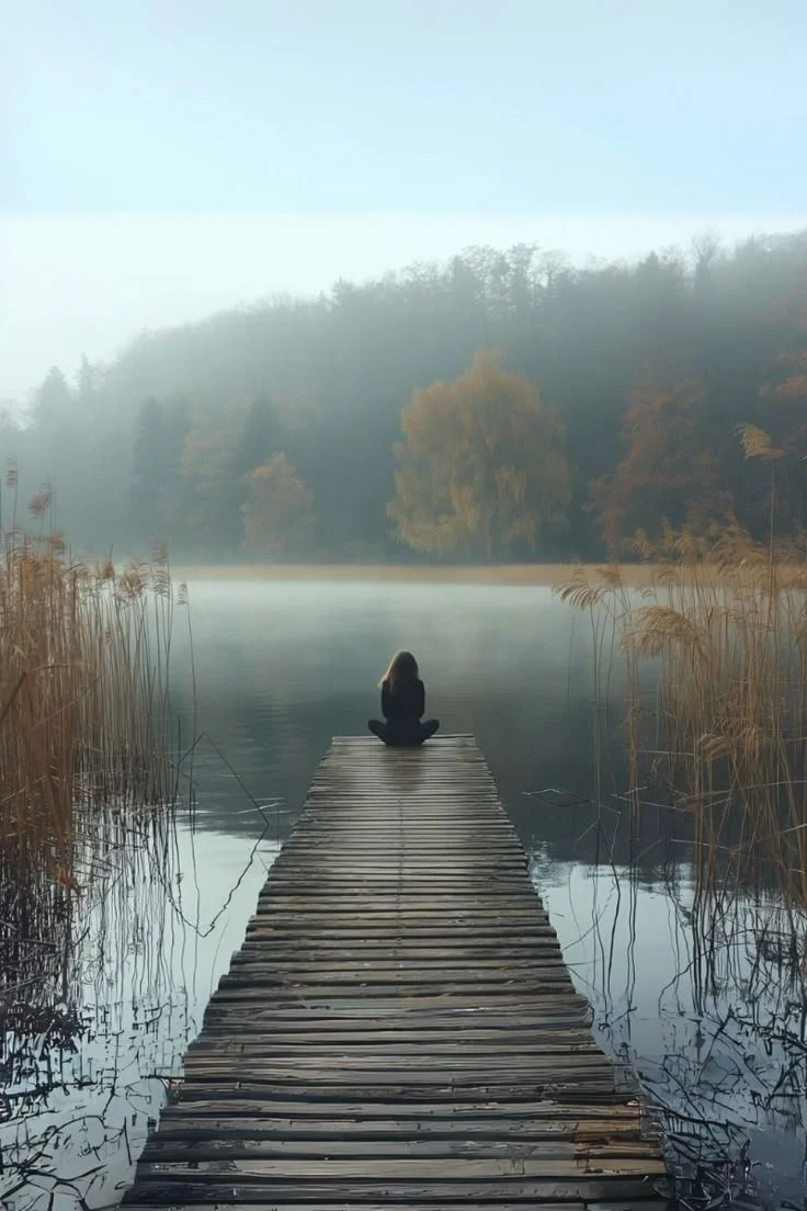 A person sitting cross-legged on a wooden dock by a calm lake, surrounded by tall grasses and trees, with fog and mist over the water and hills in the background.