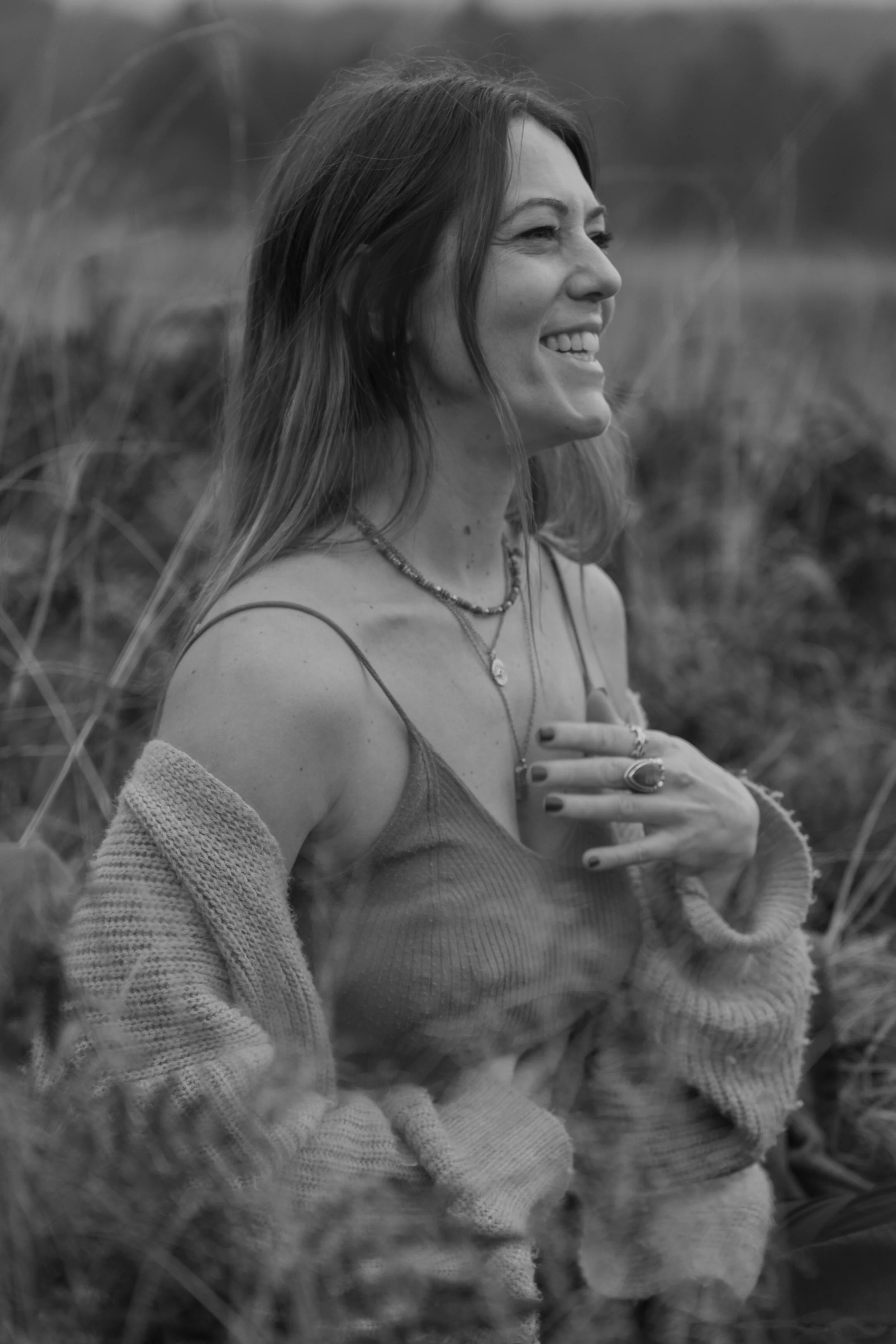 A woman smiling and looking to her right, wearing a sleeveless top and a cardigan, standing outdoors with tall grass in the background.