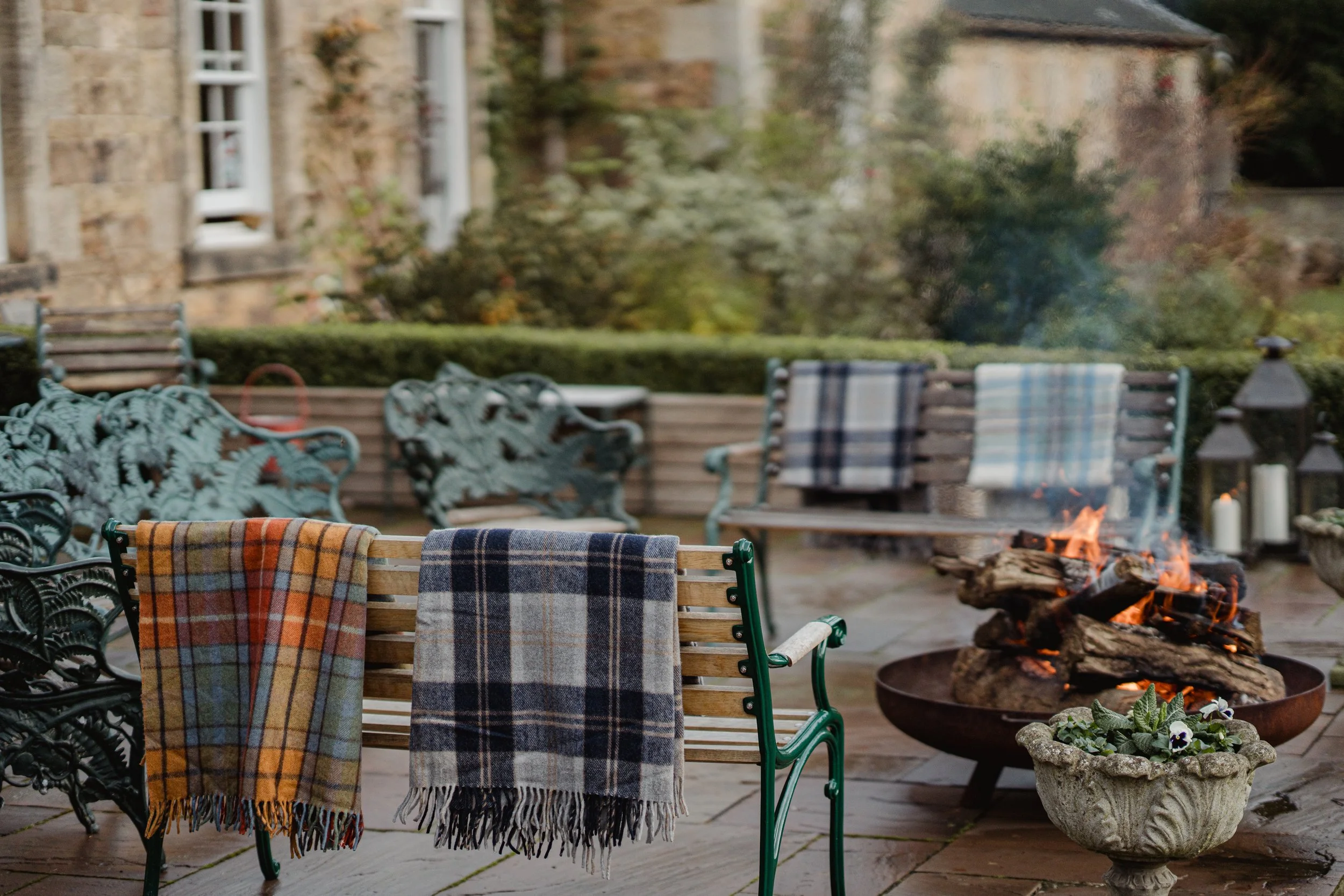 Outdoor patio with several benches, a fire pit with burning logs, and blankets draped over the benches. There are candles and lanterns in the background, and a garden with trees and shrubs.