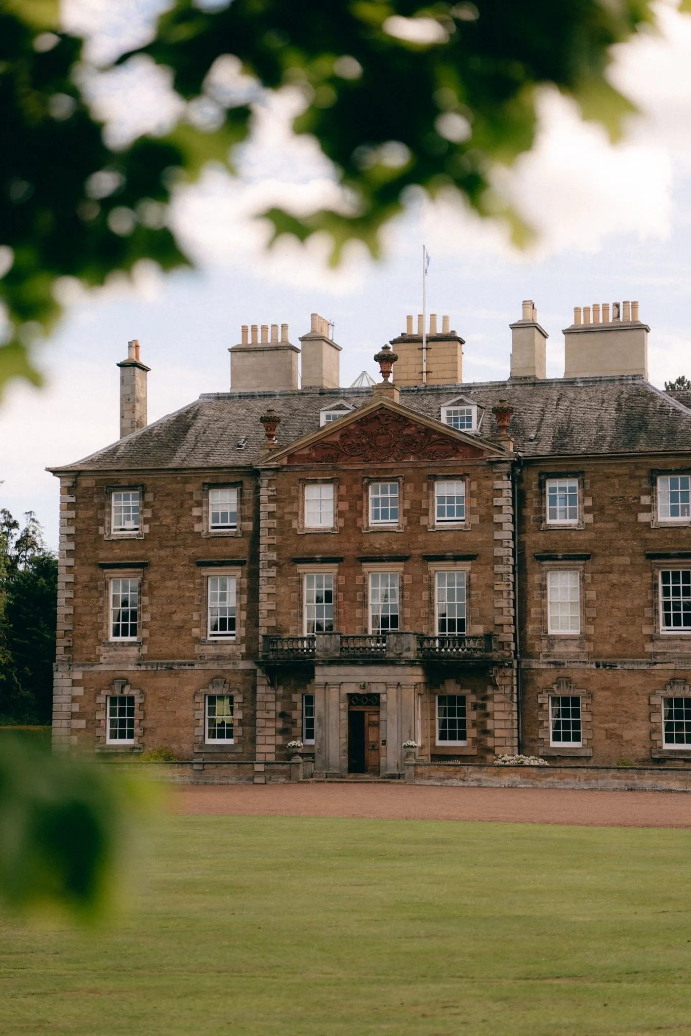 A large historic stone mansion with multiple chimneys, a mansard roof, and a flagpole on top, partially obscured by green foliage in the foreground.