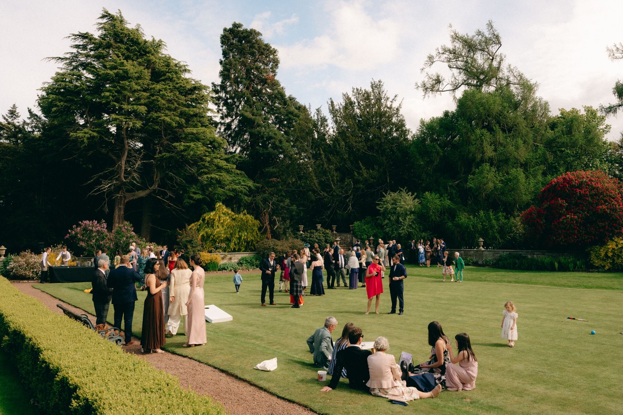 People gathered on a well-manicured lawn for an outdoor event, with large trees and a partly cloudy sky in the background.
