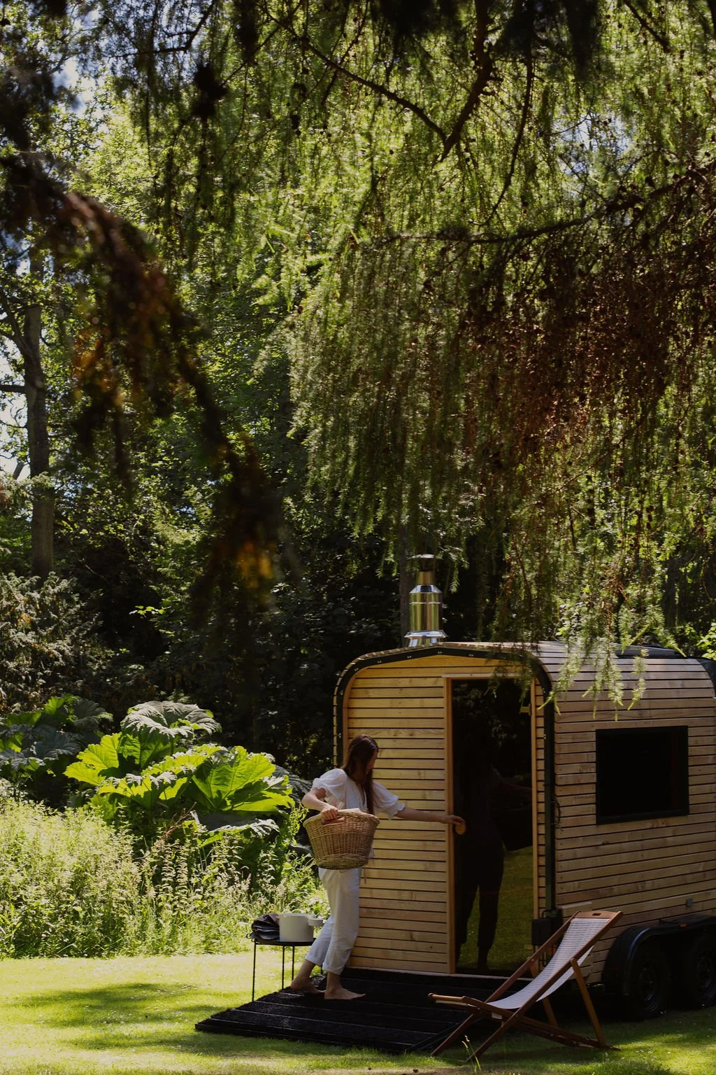 A woman in white clothes with a basket stands in front of a small wooden sauna trailer in a lush green forest, with large leafy plants nearby and a lounge chair on the grass.