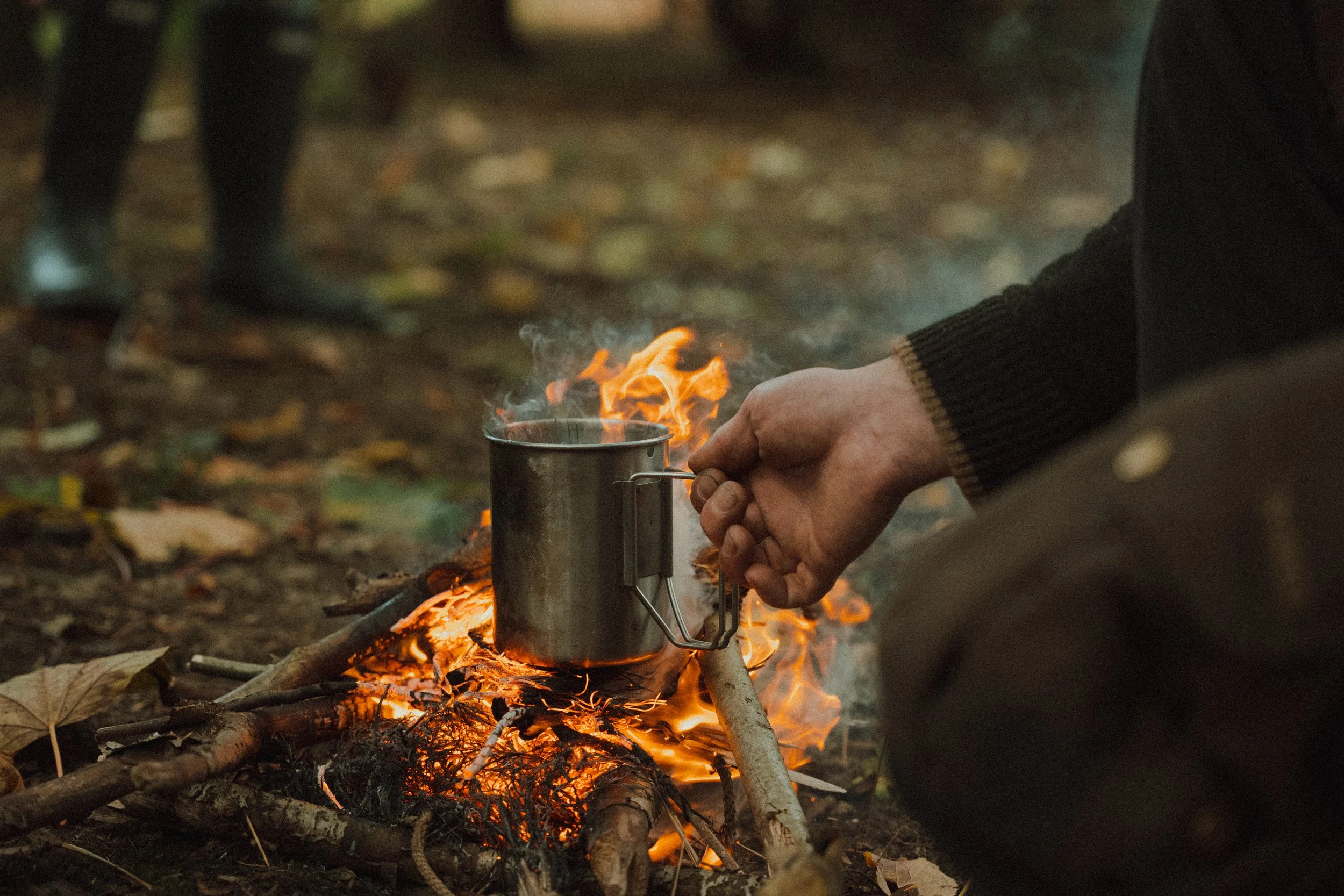 Person holding a metal cup over a campfire in an outdoor setting during autumn, with fallen leaves on the ground.