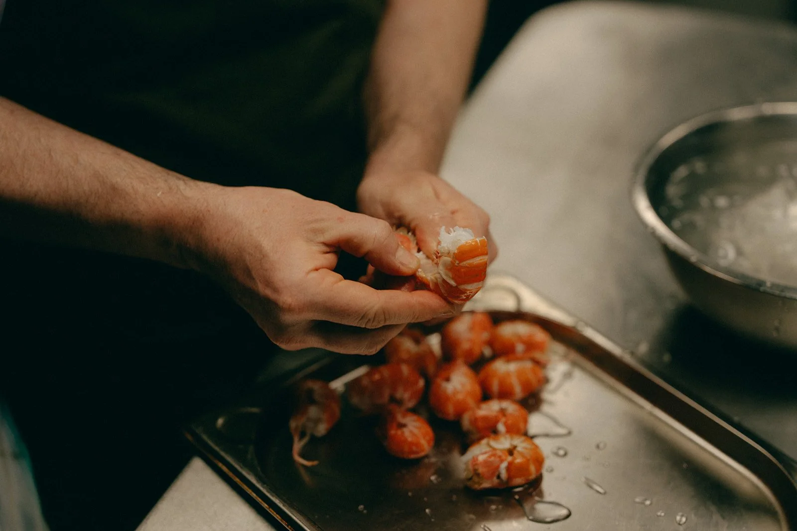 Person shelling cooked shrimp in a kitchen, with shrimp on a tray and a metal sink nearby.
