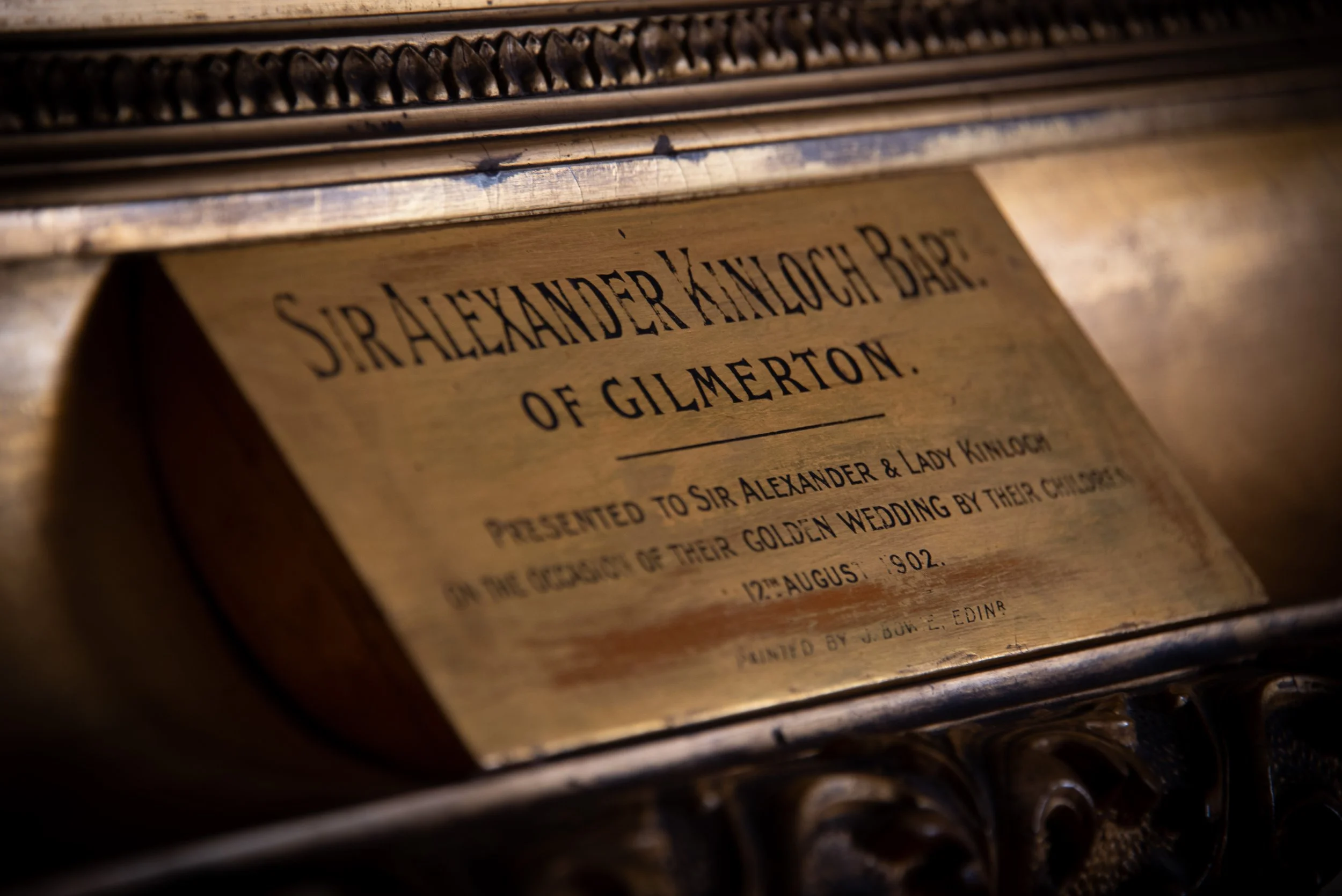 Close-up of a brass plaque with the inscription 'Sir Alexander Hamilton Baronet of Gilmerton', attached to an ornate object.