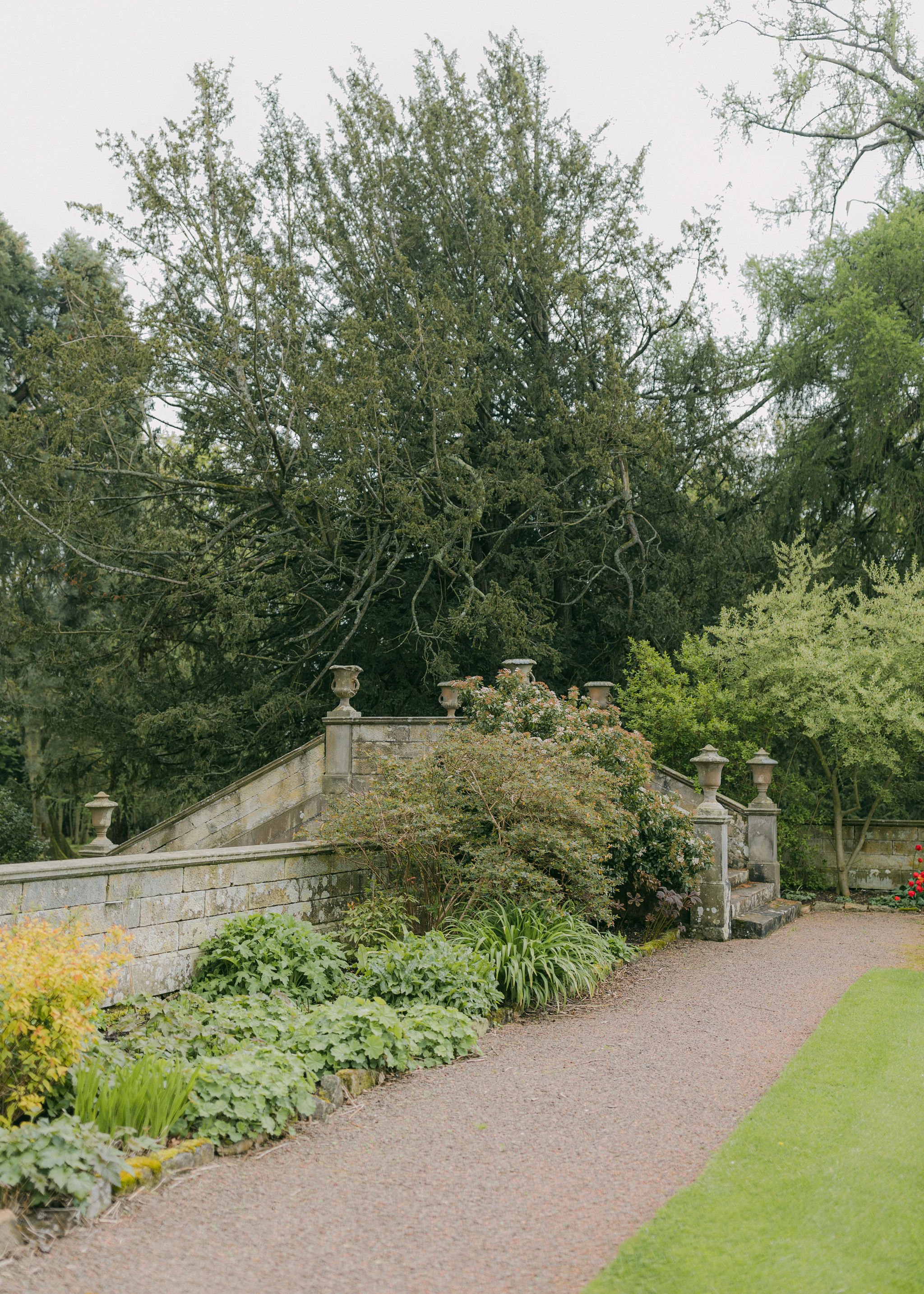 Stone staircase with decorative balustrades and urns, surrounded by lush green plants and trees in a garden setting.