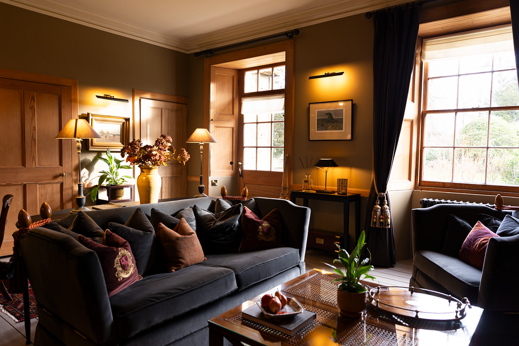 A cozy living room with wooden walls and large windows, decorated with lamps, framed pictures, a sofa with pillows, and a coffee table with a plant and decorative items.