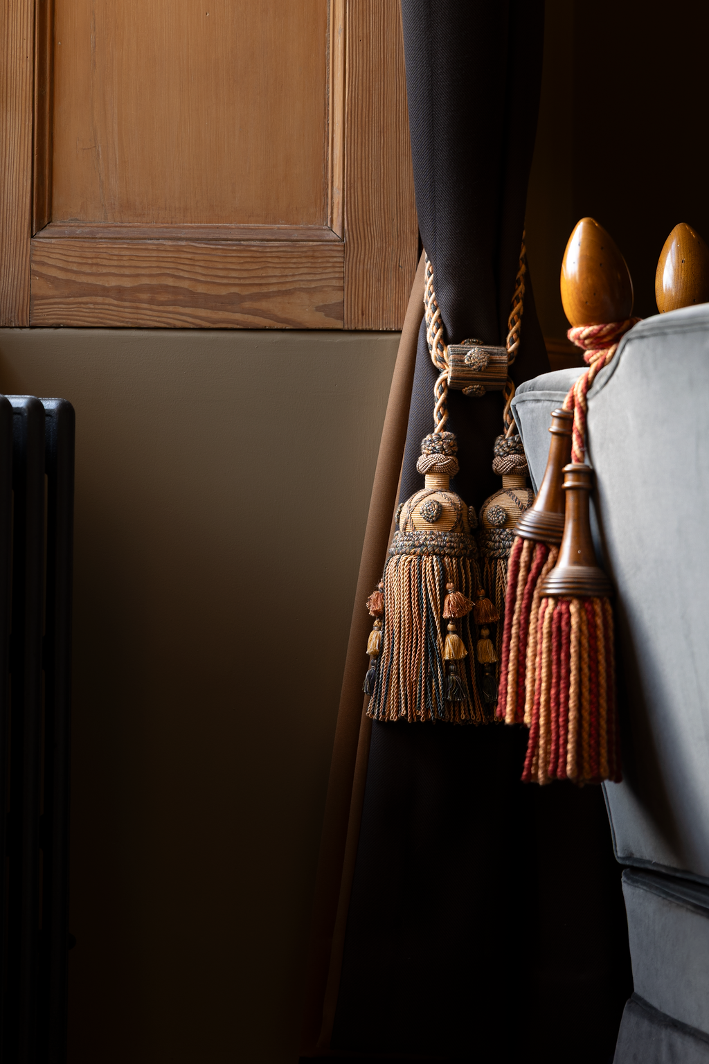 Decorative tassels hanging on a curtain next to a wooden bedpost and a grey upholstered piece of furniture.