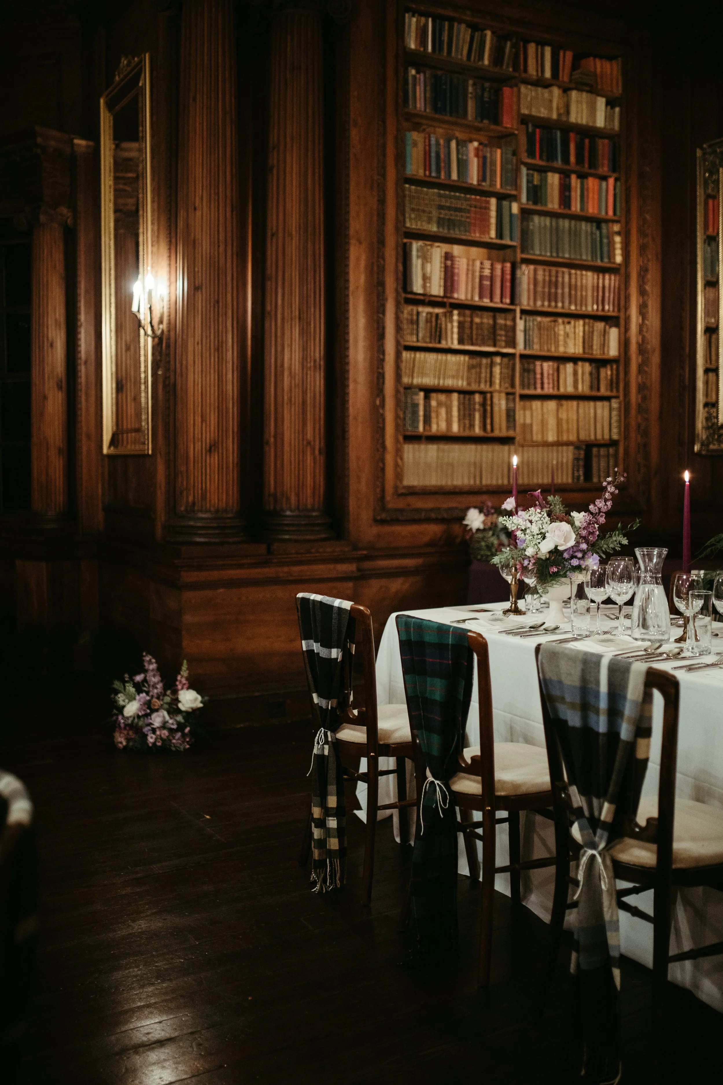 Elegant dining room with a long table set for a meal, decorated with a bouquet of flowers in a vase, purple candles, and glassware. Behind the table is a large wooden bookshelf filled with numerous books, and the walls are paneled with dark wood, creating a warm, sophisticated atmosphere.