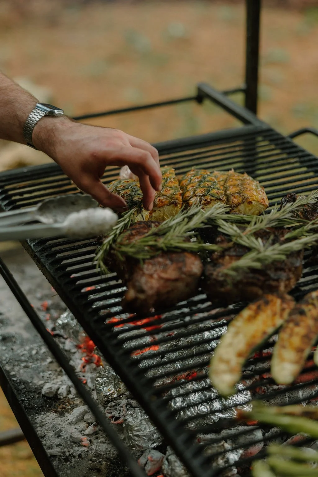 Person grilling pineapple and chicken pieces on a barbecue, garnished with rosemary.
