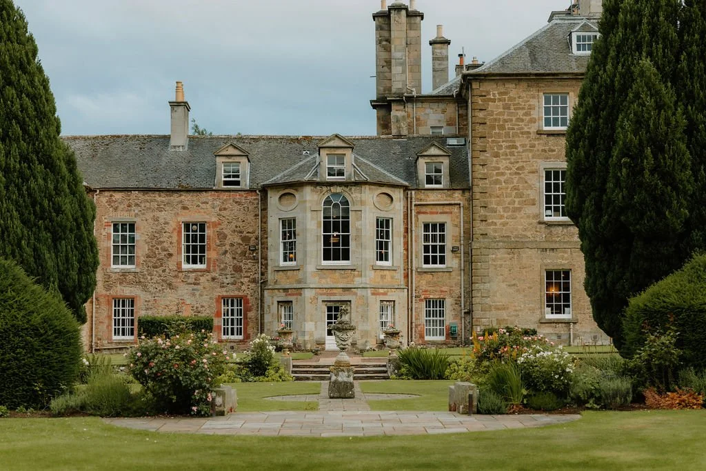 Front view of a historic stone mansion with large windows and a well-maintained garden in the foreground, featuring trimmed bushes, flowering plants, and a stone pathway.