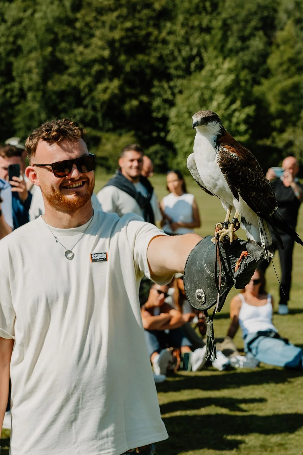A man wearing sunglasses and a white t-shirt smiling while holding a falcon on his gloved arm in a park with several people in the background.