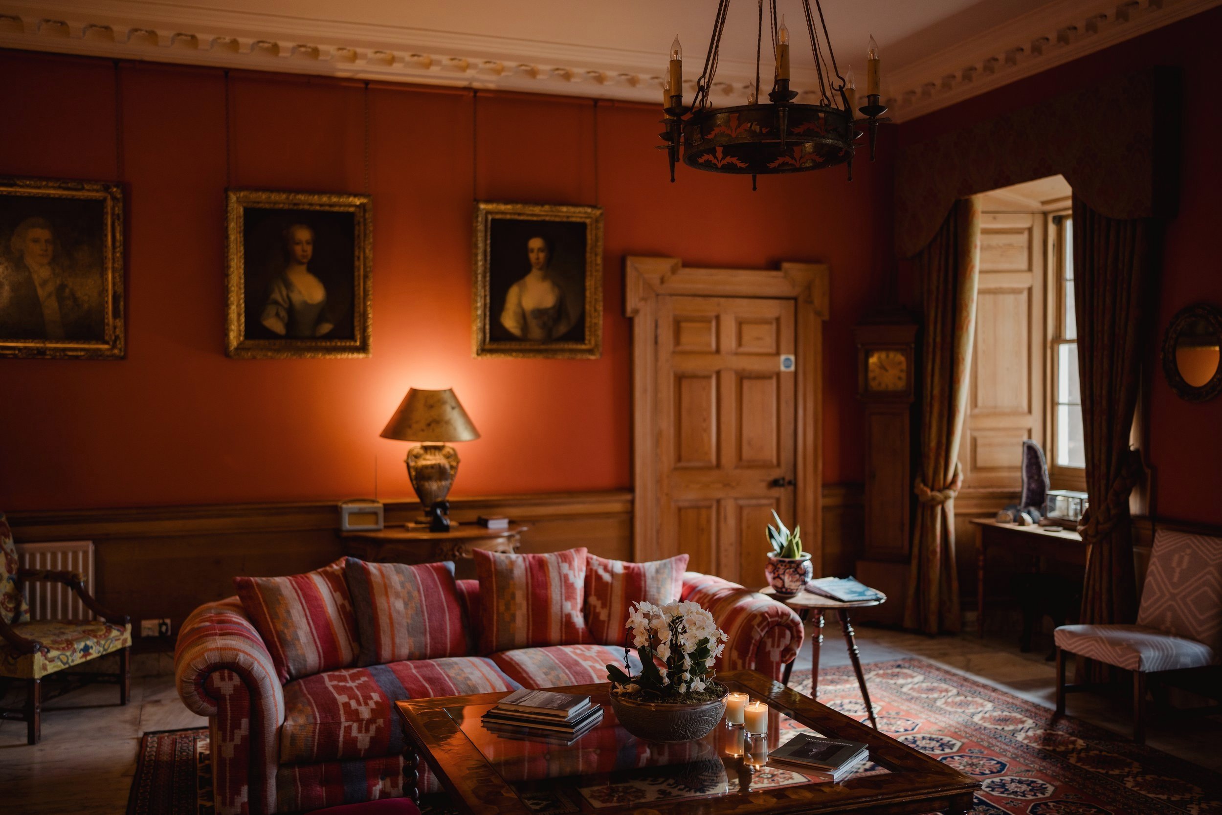 A cozy living room with a red wall, framed portraits, a wooden door, and a window with heavy curtains. There is a patterned sofa, a coffee table with a flower arrangement and candles, and a lamp emitting warm light.