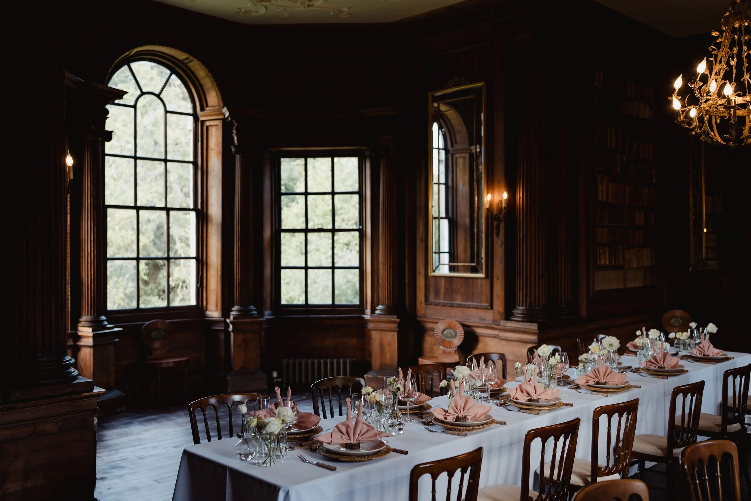 Elegant dining room with large arched windows, dark wood paneling, a chandelier, and a table set for a formal meal with pink napkins and floral centerpieces.