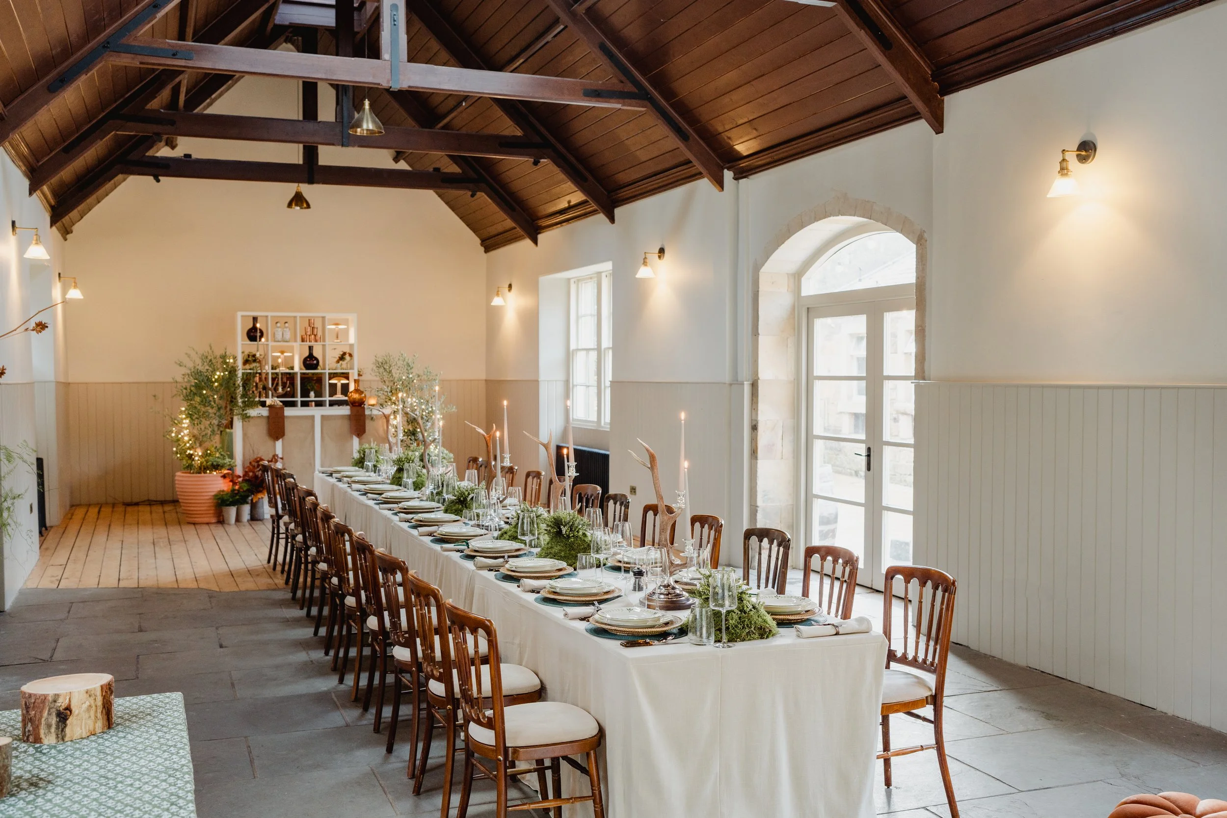 Warmly lit dining room with a long table set for a meal, decorated with greenery and candles. The room has a high wooden ceiling with exposed beams, large windows, and simple wall-mounted lamps.