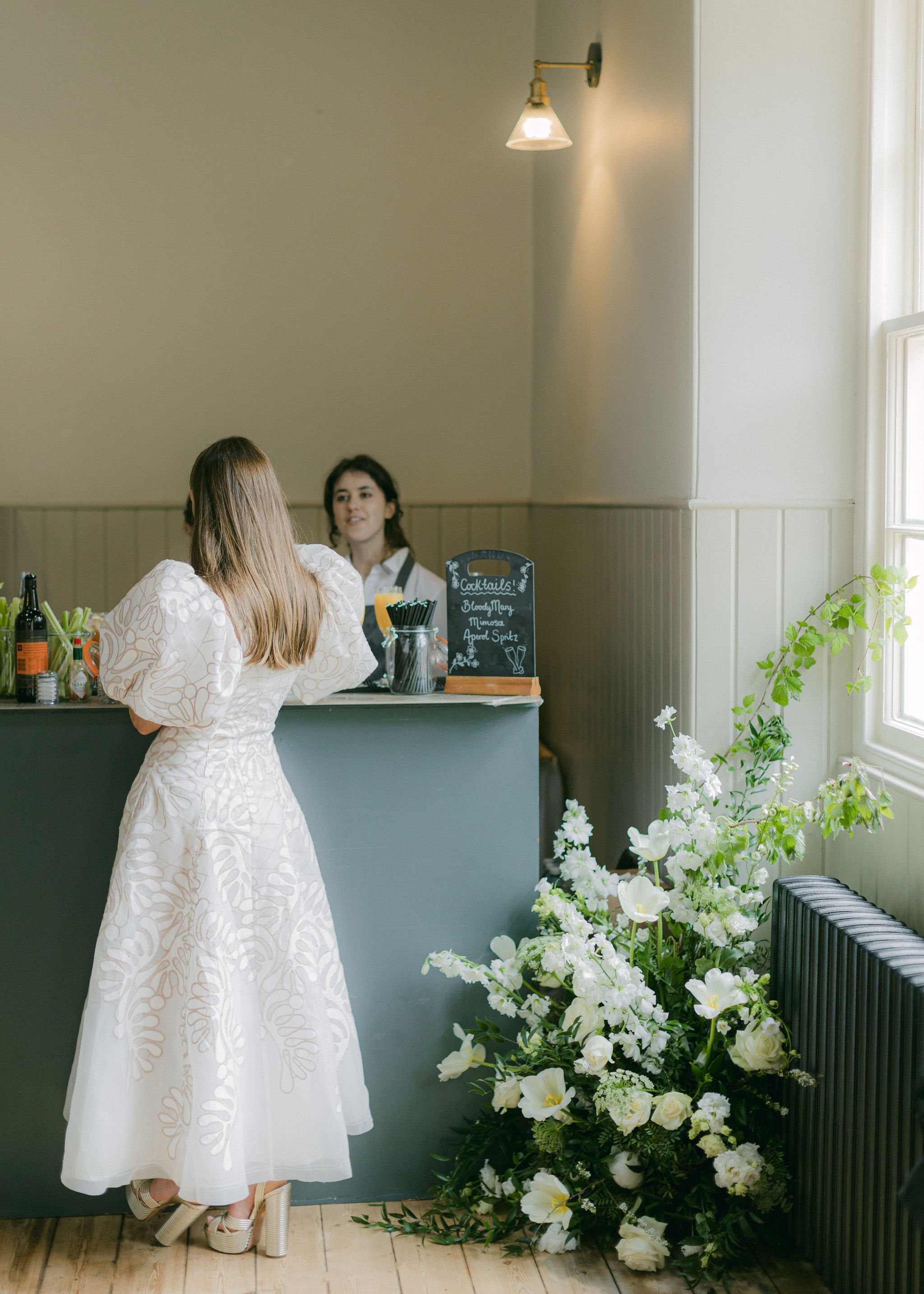 A woman in a white dress with beige patterns is standing indoors near a counter, facing away from the camera, wearing high-heeled shoes. Behind the counter, a bartender with shoulder-length dark hair, wearing a white shirt and black apron, is smiling. The counter has various bottles and a small chalkboard menu listing cocktails such as Bloody Mary, Mimosa, and Aperol Spritz. To the right, a large bouquet of white flowers and greenery is placed next to a heat radiator near a window with light streaming in.