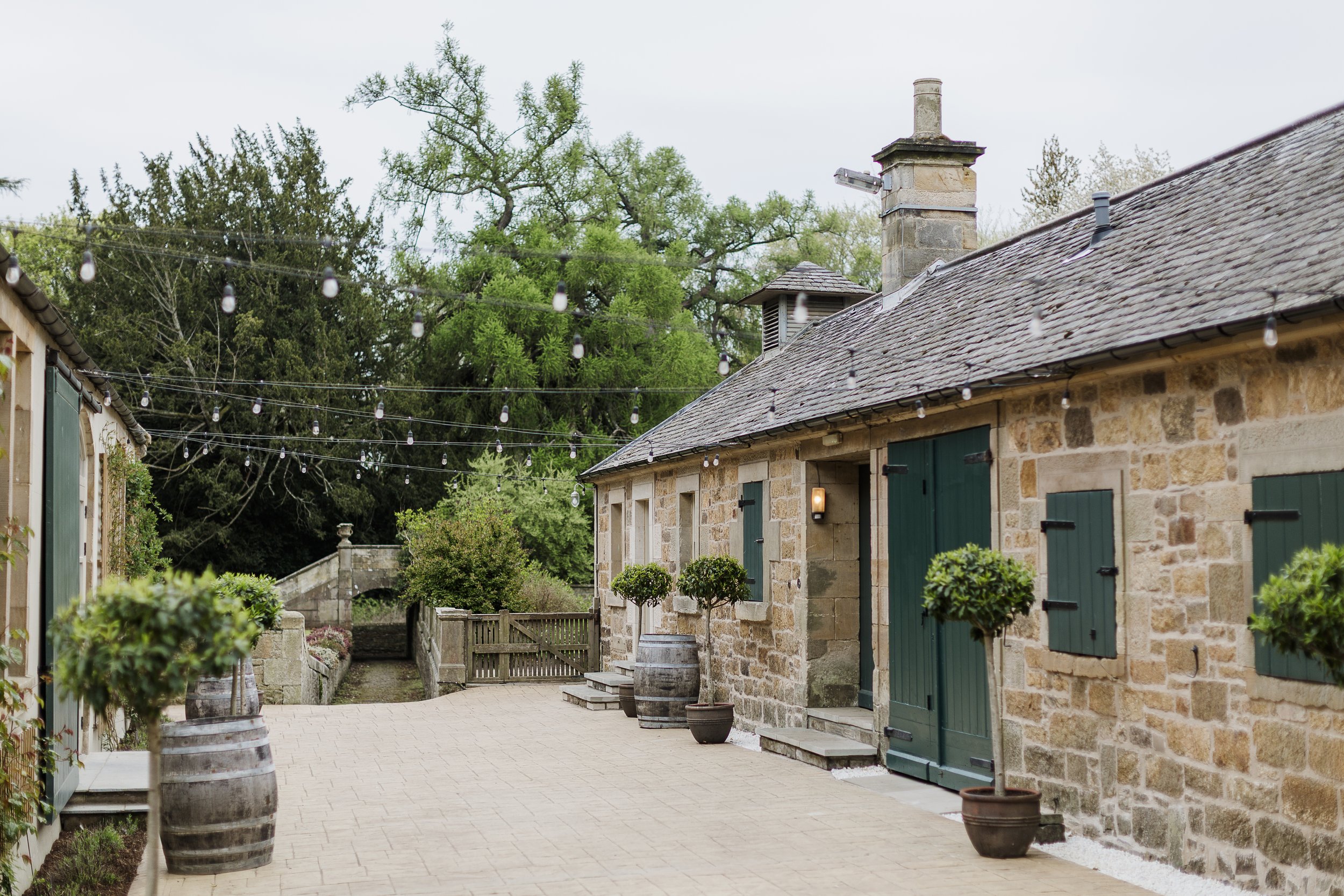 A rustic courtyard with stone buildings, green shutters, potted plants, and string lights, surrounded by trees.