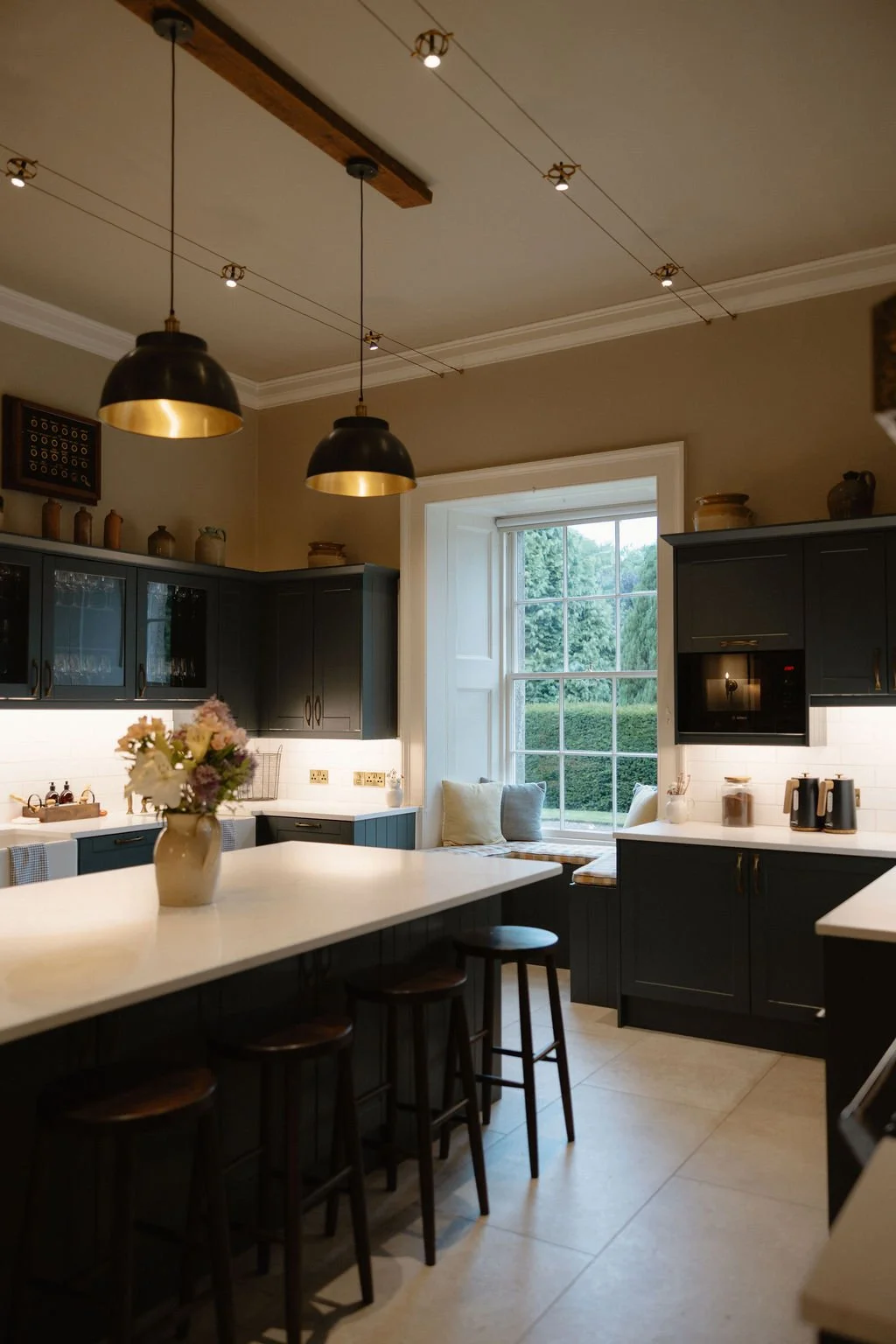 Modern kitchen with dark cabinets, white countertops, a large window with a cushioned bench, and hanging black pendant lights with gold interiors.
