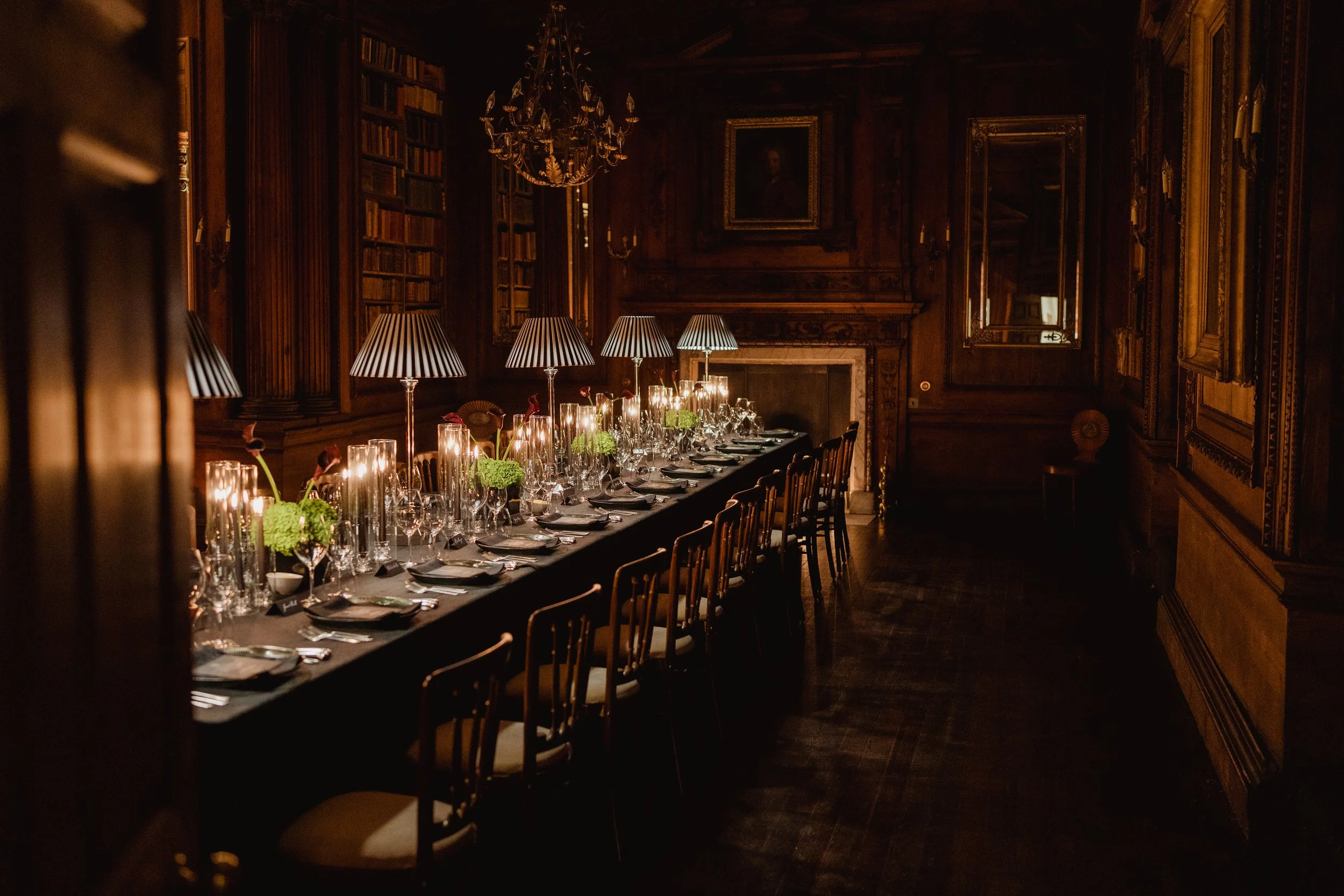 A dimly lit, elegant dining room with a long table adorned with candles, glassware, and floral arrangements, surrounded by wooden chairs, with bookshelves, mirrors, a chandelier, and a portrait hanging on the dark wood-paneled walls.
