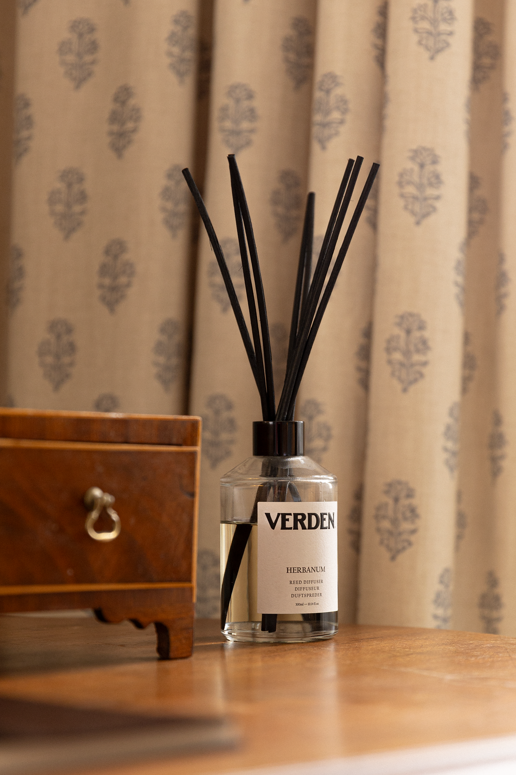 A reed diffuser with black reeds in a clear glass bottle labeled 'VERDEN HERBANUM' on a wooden tabletop, with a wooden drawer and a beige curtain with blue floral pattern in the background.