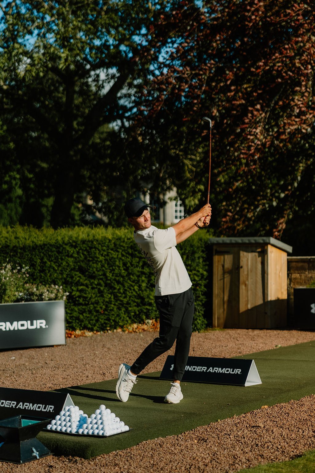 A man practicing golf, swinging a club on a mat at an outdoor driving range during daytime, with trees and a wooden structure in the background.