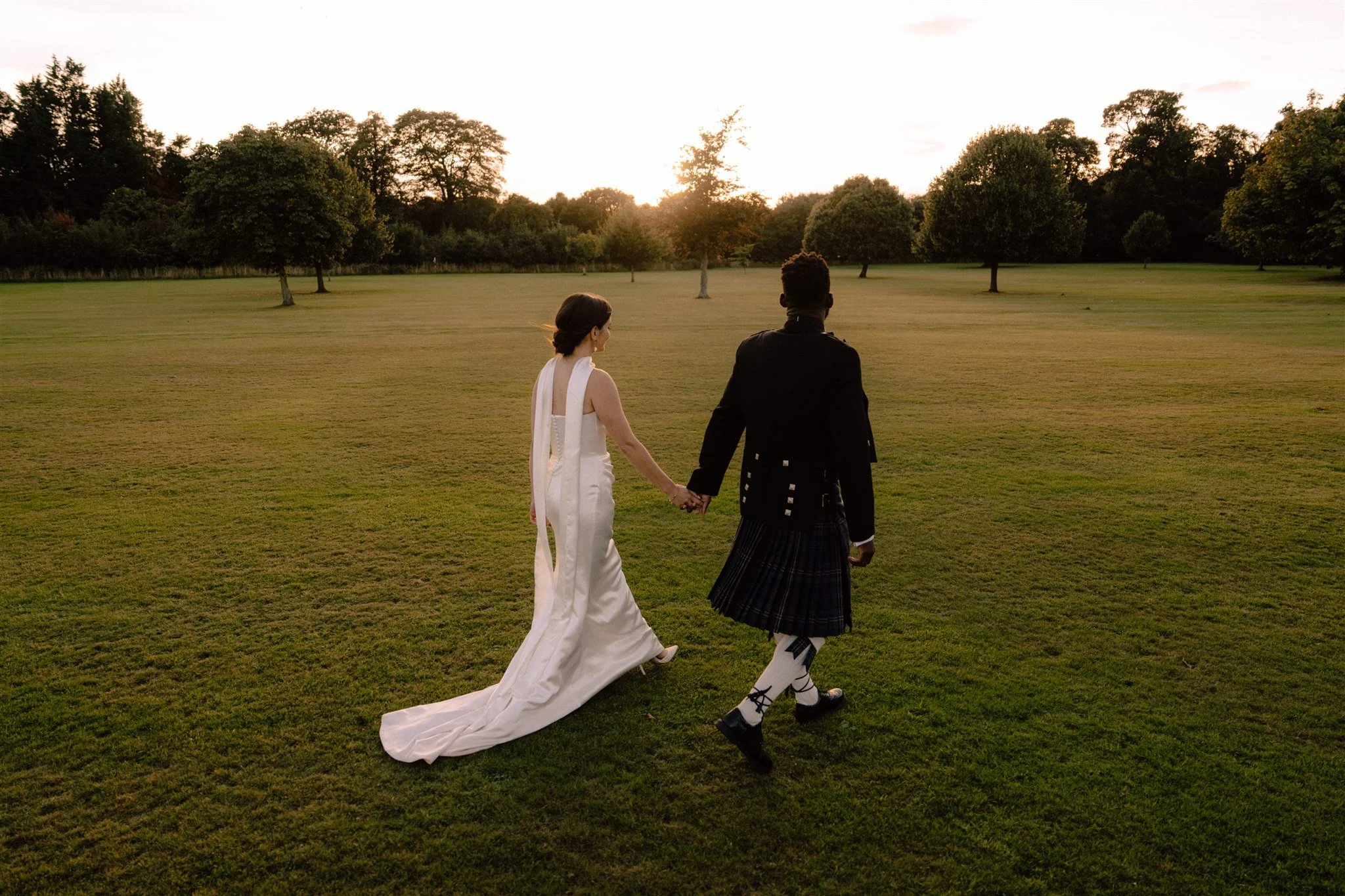 A couple holding hands and walking across a grassy field at sunset, with trees in the background.