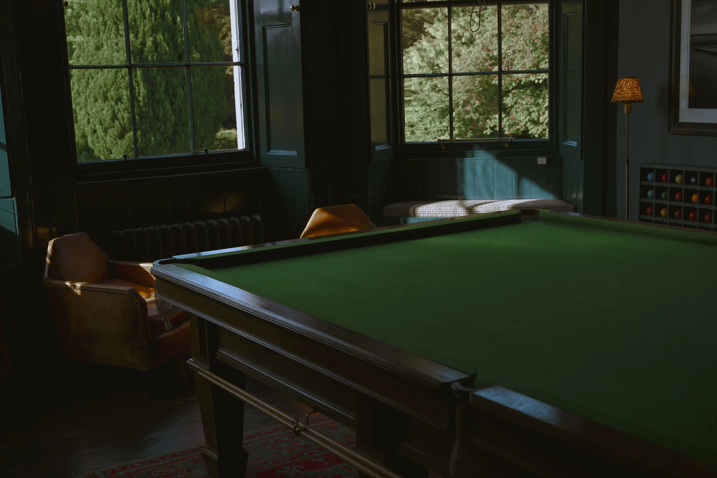 Interior of a room with a green billiards table, two brown chairs, large windows showing green trees outside, a lamp, and a wall with a framed picture and a colorful ball rack.
