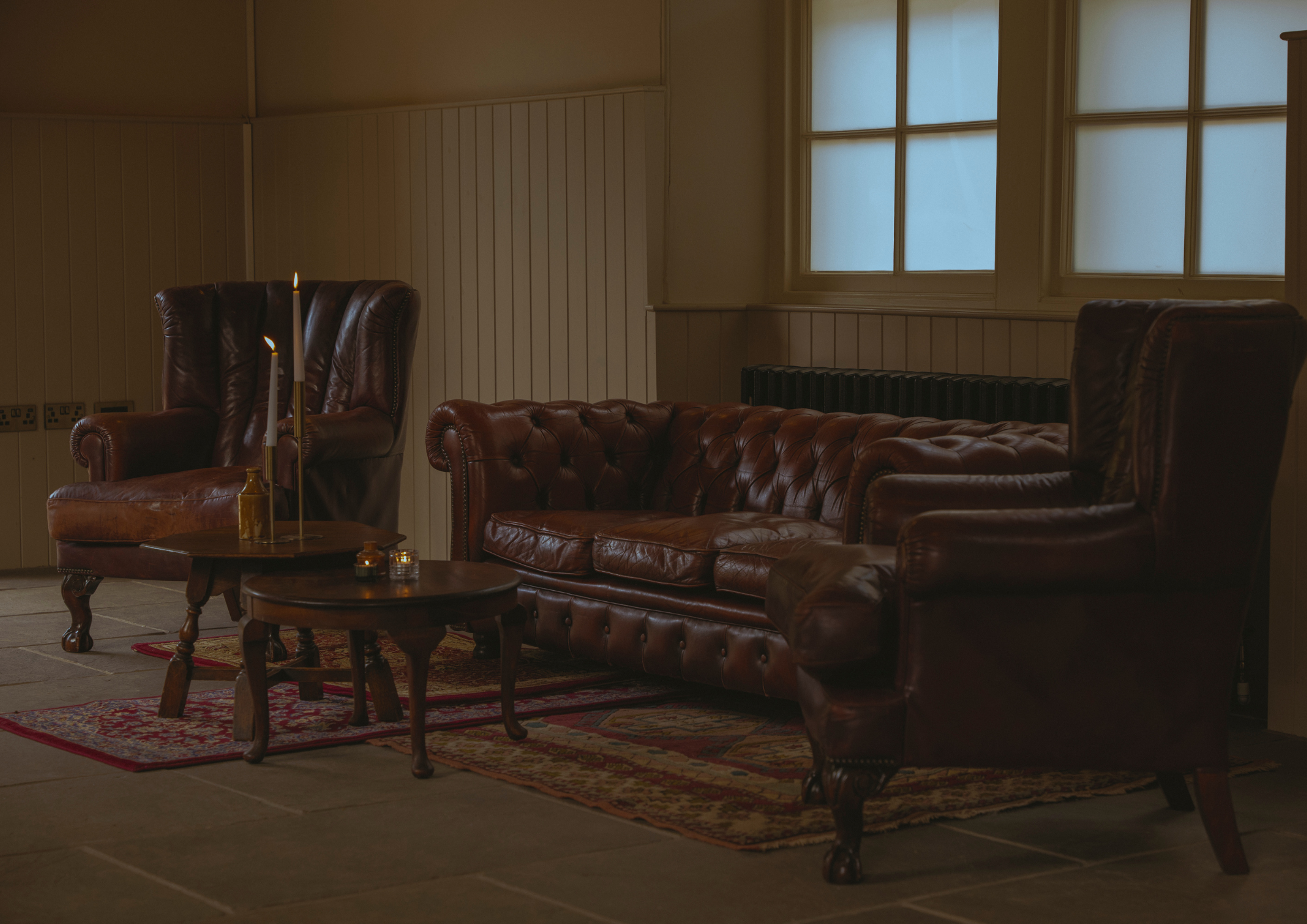 A cozy living room corner with two brown leather armchairs, a matching sofa, wooden coffee tables, a decorative rug, and large windows letting in natural light.