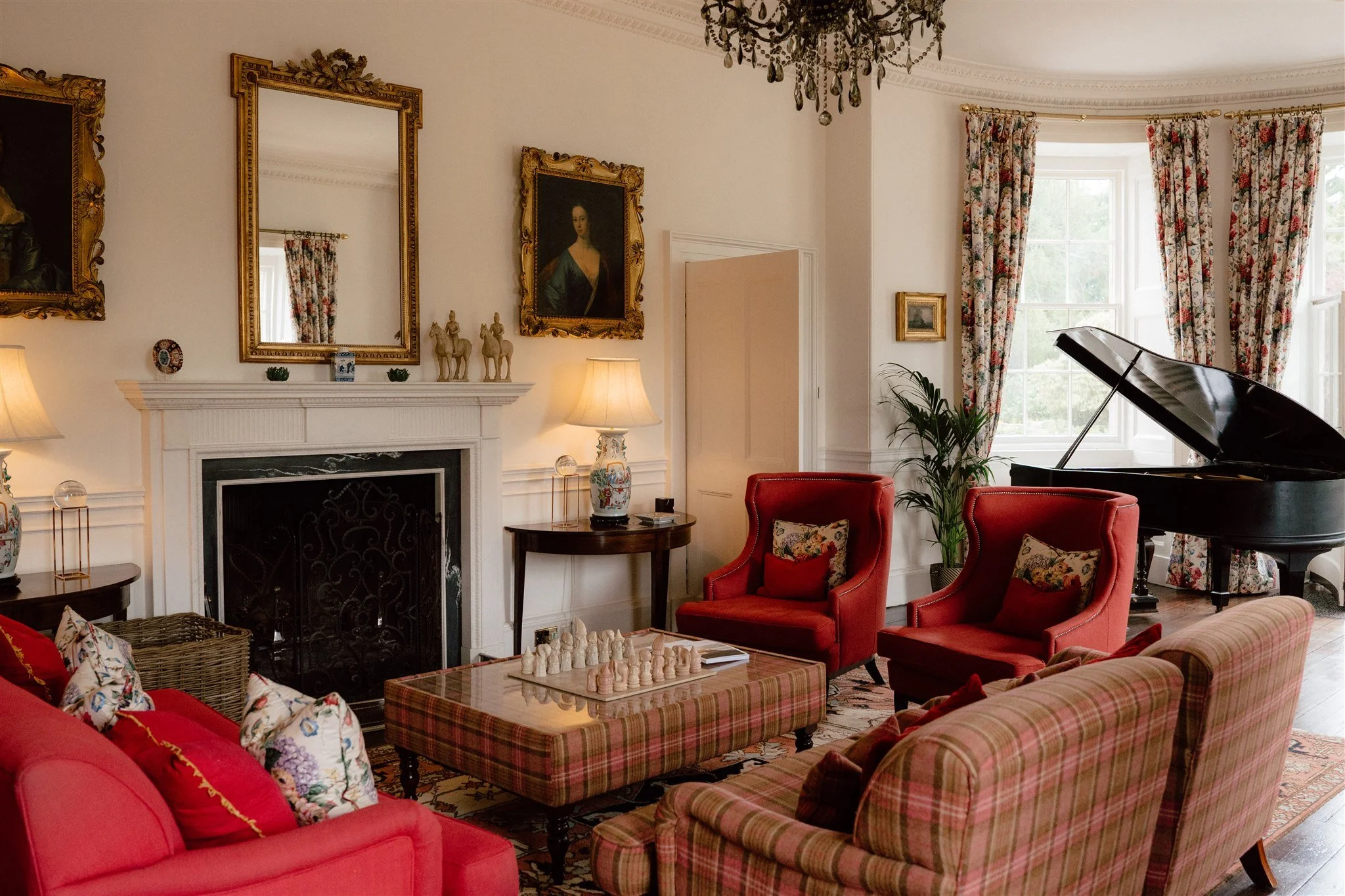 A cozy, elegant living room with a white fireplace and black marble insert, decorated with ornate gold-framed mirrors and paintings, floral curtains framing large windows, and red upholstered chairs with floral pillows. A black grand piano is positioned near the windows, and there are various lamps and decorative items throughout the room.