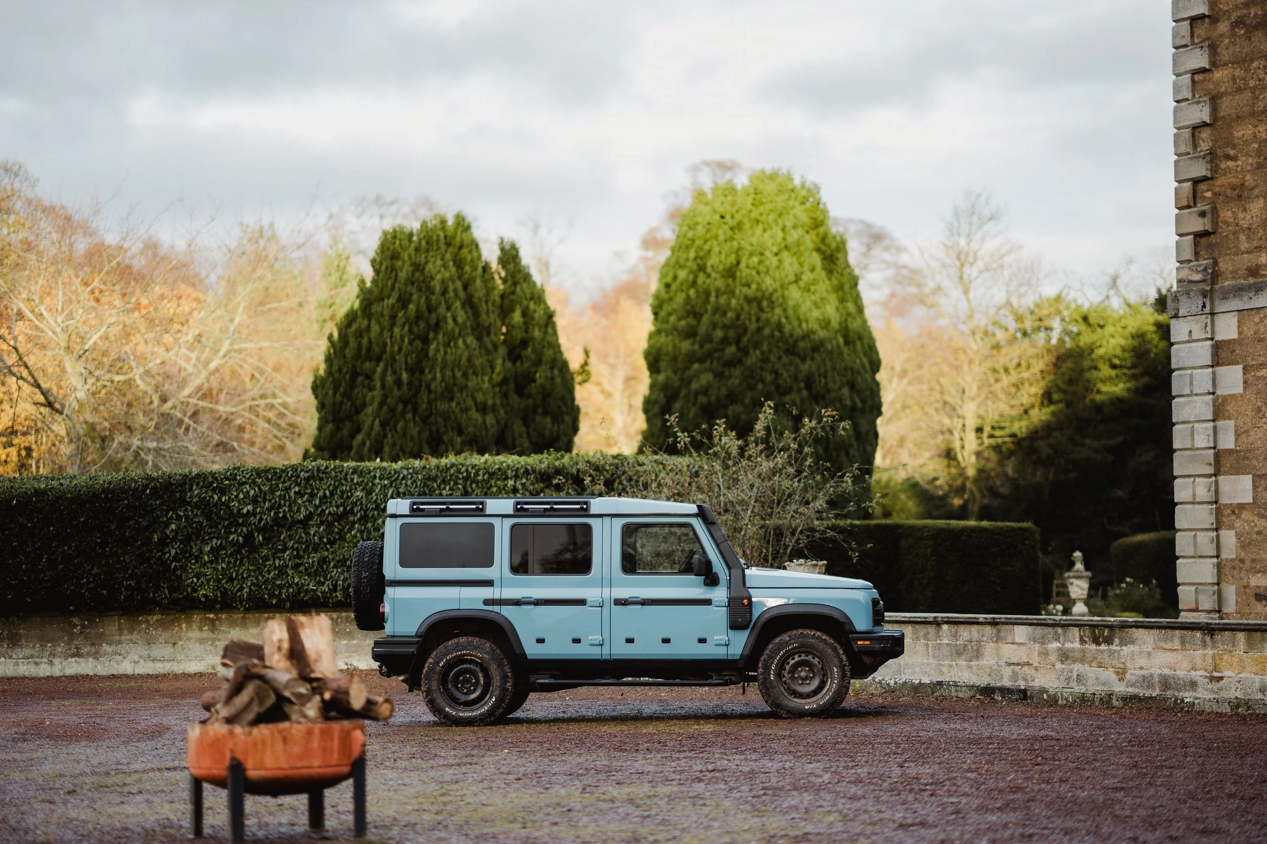 A light blue Toyota Land Cruiser parked on a paved area near a stone wall, with a garden of tall green trees and bushes in the background and cloudy sky overhead.