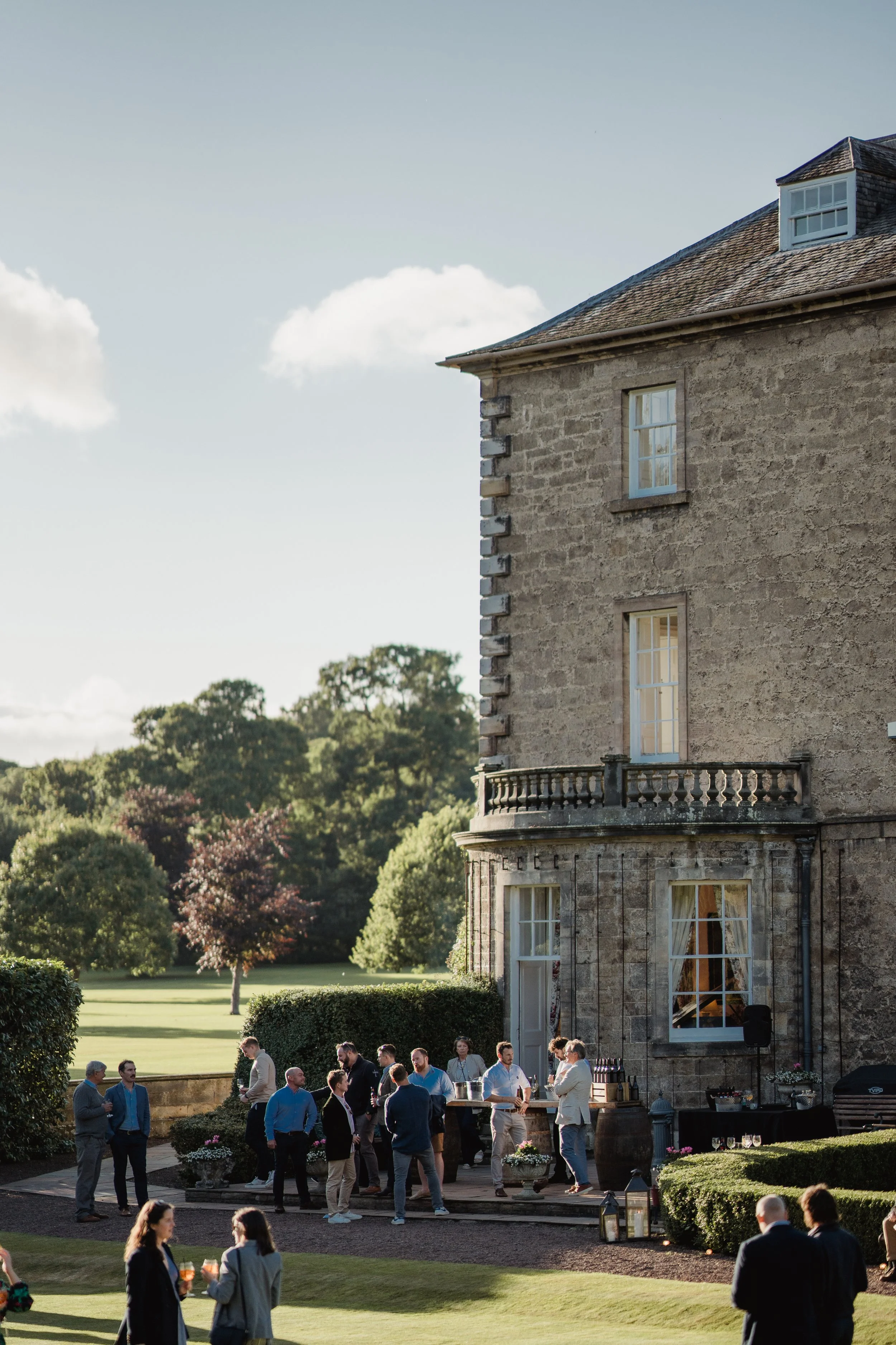 People gathering outdoors for a social event near a stone building with large windows, on a lawn with trees in the background during daytime.