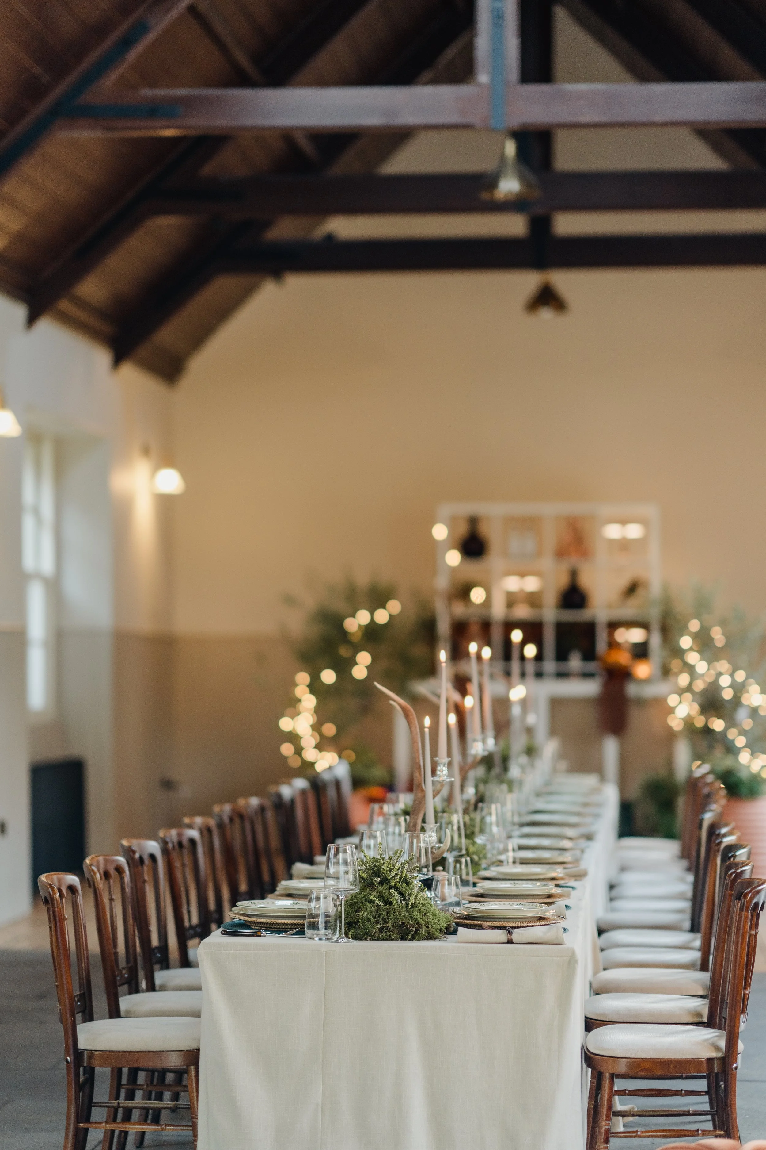 A long dining table set for a formal event in a venue with high ceilings and exposed beams, decorated with greenery and candles, with chairs on both sides and blurred background of warm lighting and wall decorations.
