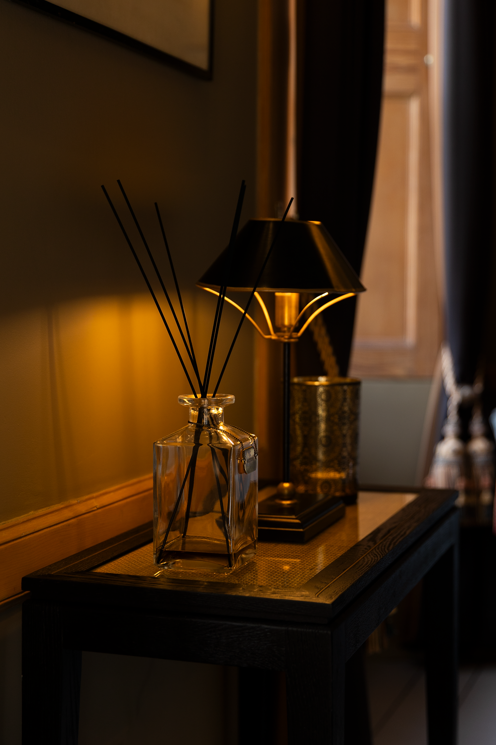 Decorative glass bottle with black reed diffuser sticks and a black table lamp on a wooden table, with a gold-patterned container and a wooden cabinet in the background.