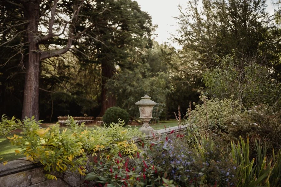 A stone garden wall with an ornate stone garden lamp and lush plants and trees in the background.