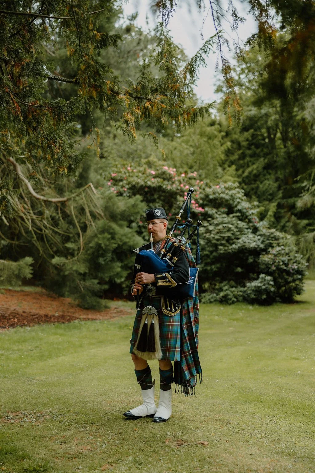 A man dressed in traditional Scottish attire playing bagpipes outdoors in a lush garden with trees and shrubs.