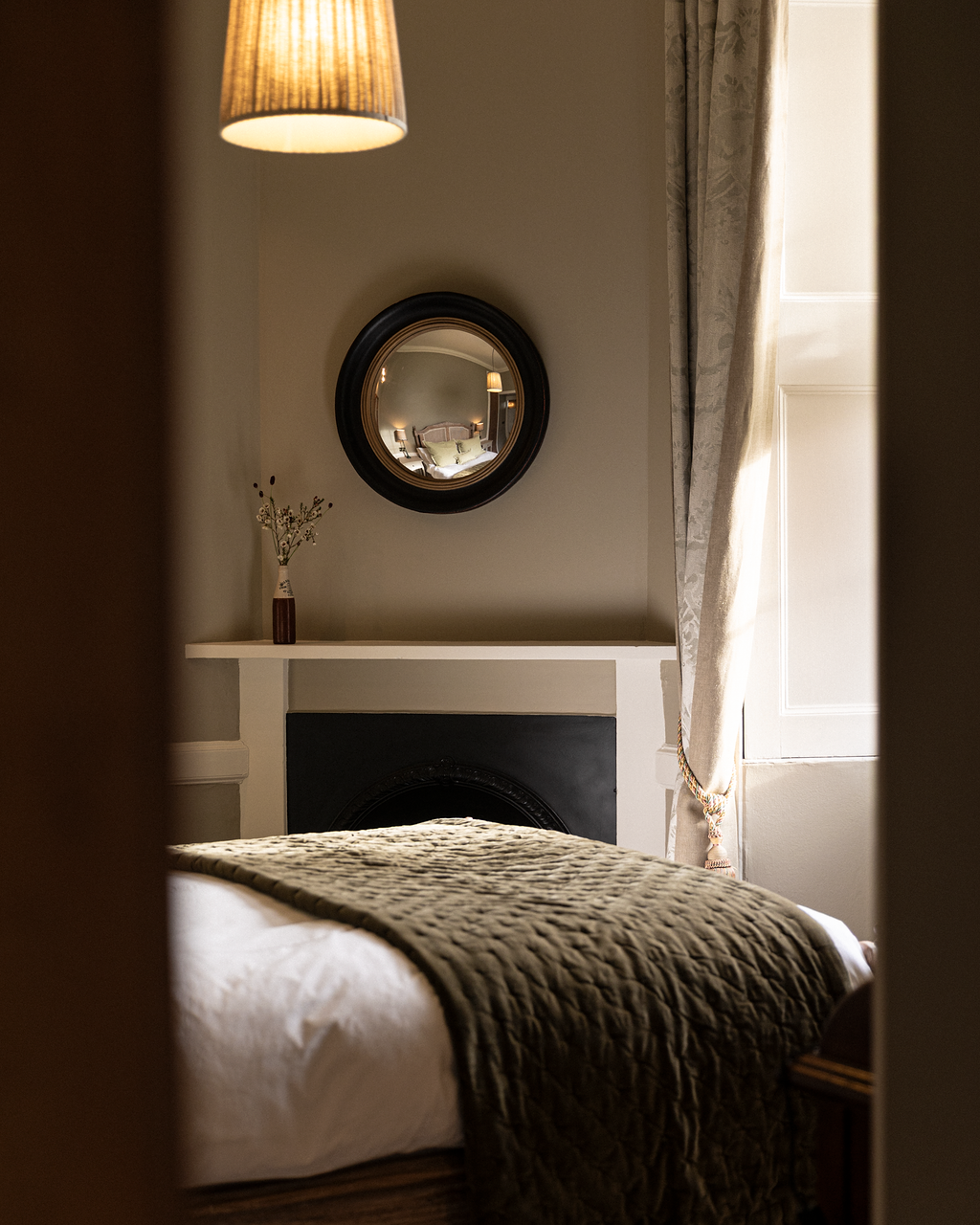 A cozy bedroom seen through a partially opened door, featuring a bed with white sheets and a textured olive-green blanket, a round mirror on the wall reflecting the bed and bedside lamps, a small vase with simple flower decorations on a ledge, beige curtains, and warm lighting from a hanging lamp.